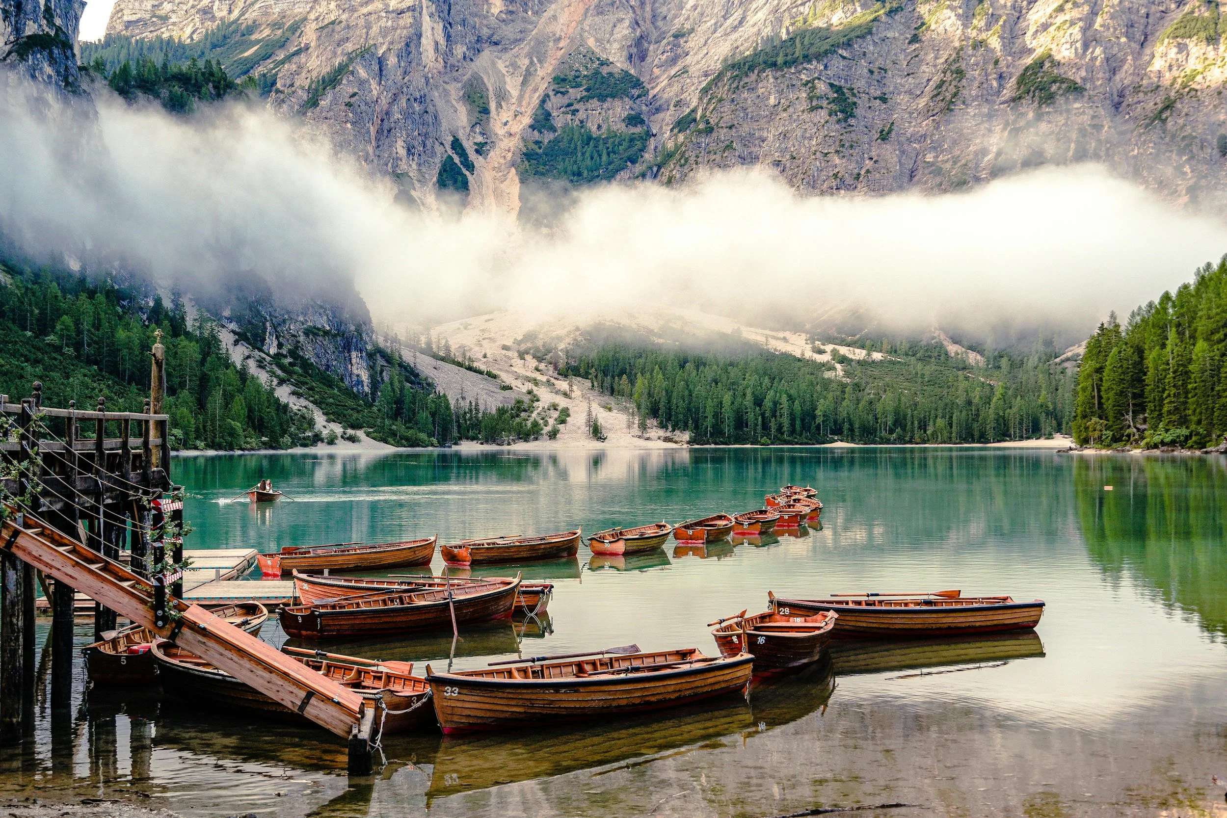 A scenic mountain lake with wooden boats docked at a pier, surrounded by green trees and a misty mountain backdrop.