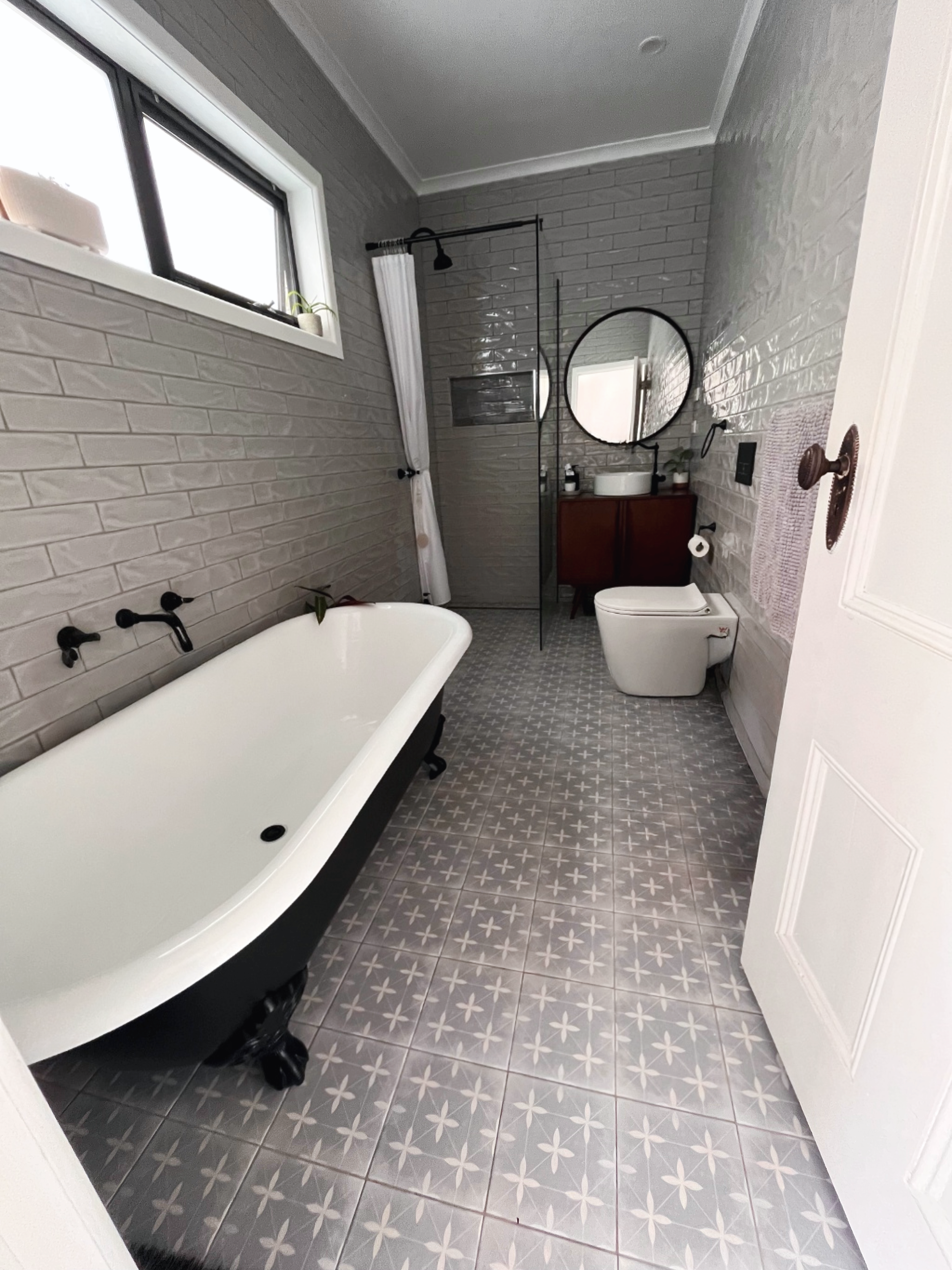 A modern bathroom with a clawfoot bathtub, a toilet, a wooden vanity with a round mirror, a shower with a white curtain, a small window with a plant on the windowsill, and gray tiled walls and flooring with a white star pattern.