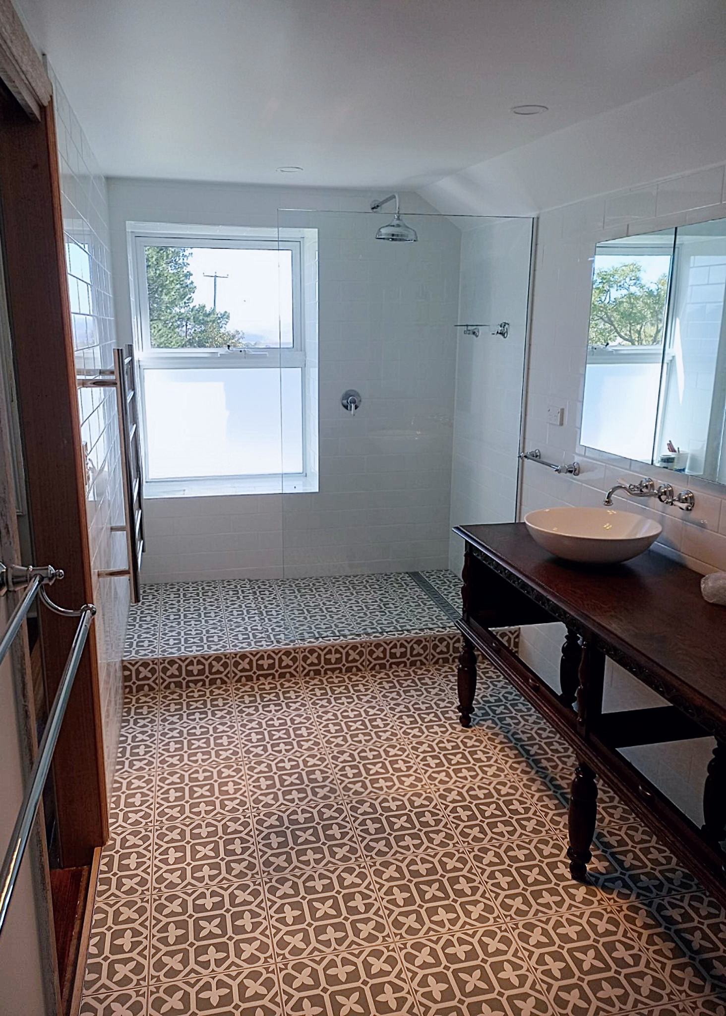 Bathroom with patterned floor tiles, a window, glass shower with silver fixtures, a mirror, and a wooden vanity with a vessel sink.