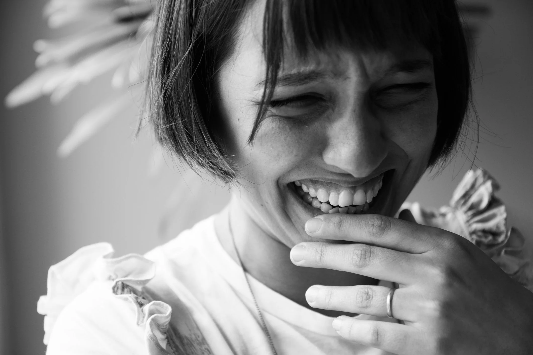 Black and white close-up of a woman laughing with her eyes closed, hand near her mouth, short hair, wearing a ring and a ruffled top.