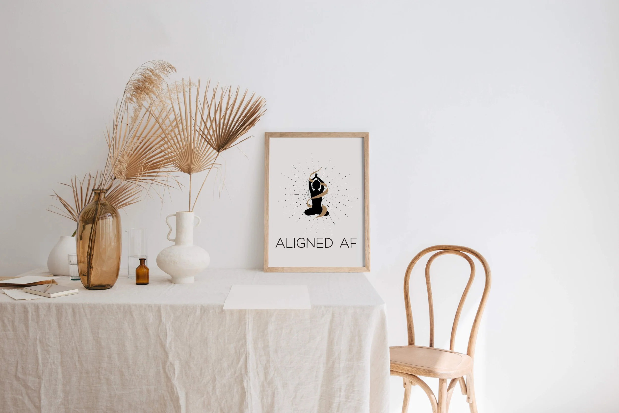 A framed print reading "ALIGNED AF" sits on a table with dried plants in vases and a wooden chair against a plain white wall.