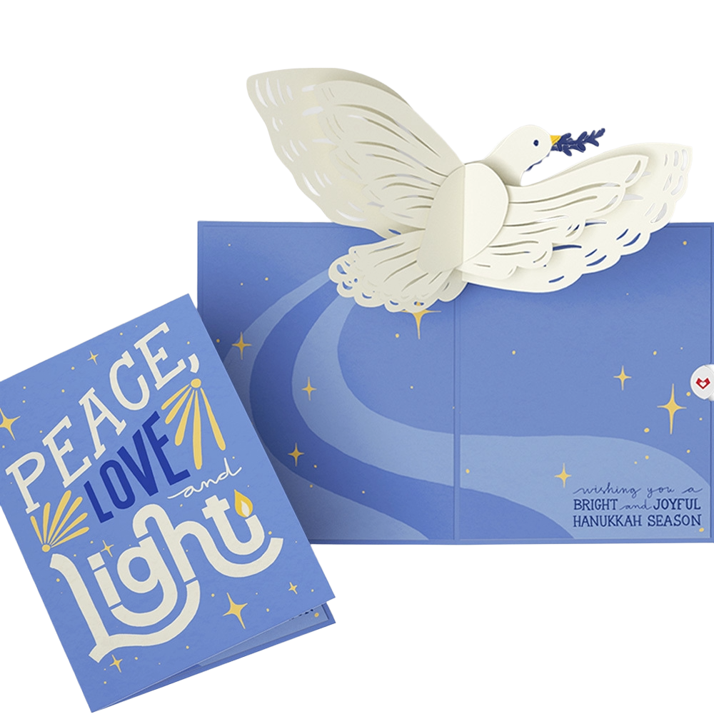 A Hanukkah greeting card with a dove carrying an olive branch, sitting on a blue box decorated with a moon and stars. The card says 'Peace, Love and Light' and the box has a message wishing a bright and joyful Hanukkah season.