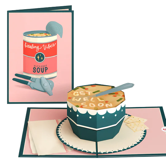 A birthday cake with a 'Get Well Soon' message, a knife on top, and two crackers next to it, in front of a pink card with a soup can illustration and the words 'Healing Vibes'.