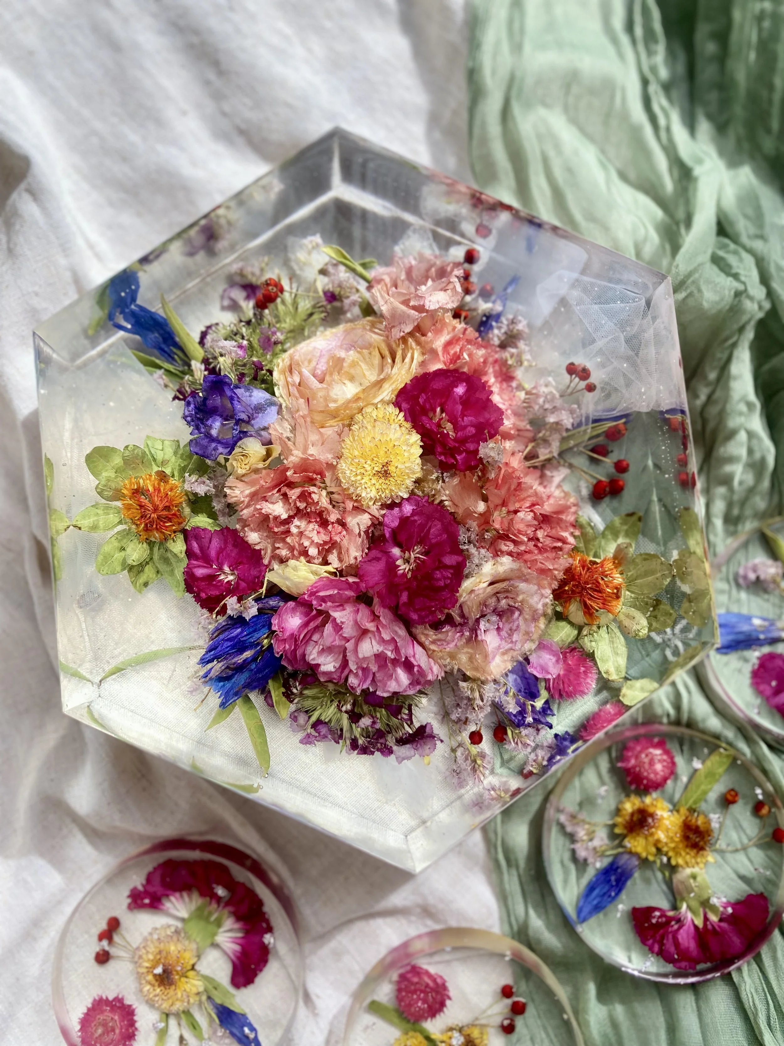 Colorful bouquet of fresh flowers wrapped in clear paper and displayed on a table, alongside smaller floral arrangements in glass bowls, with a green fabric backdrop.