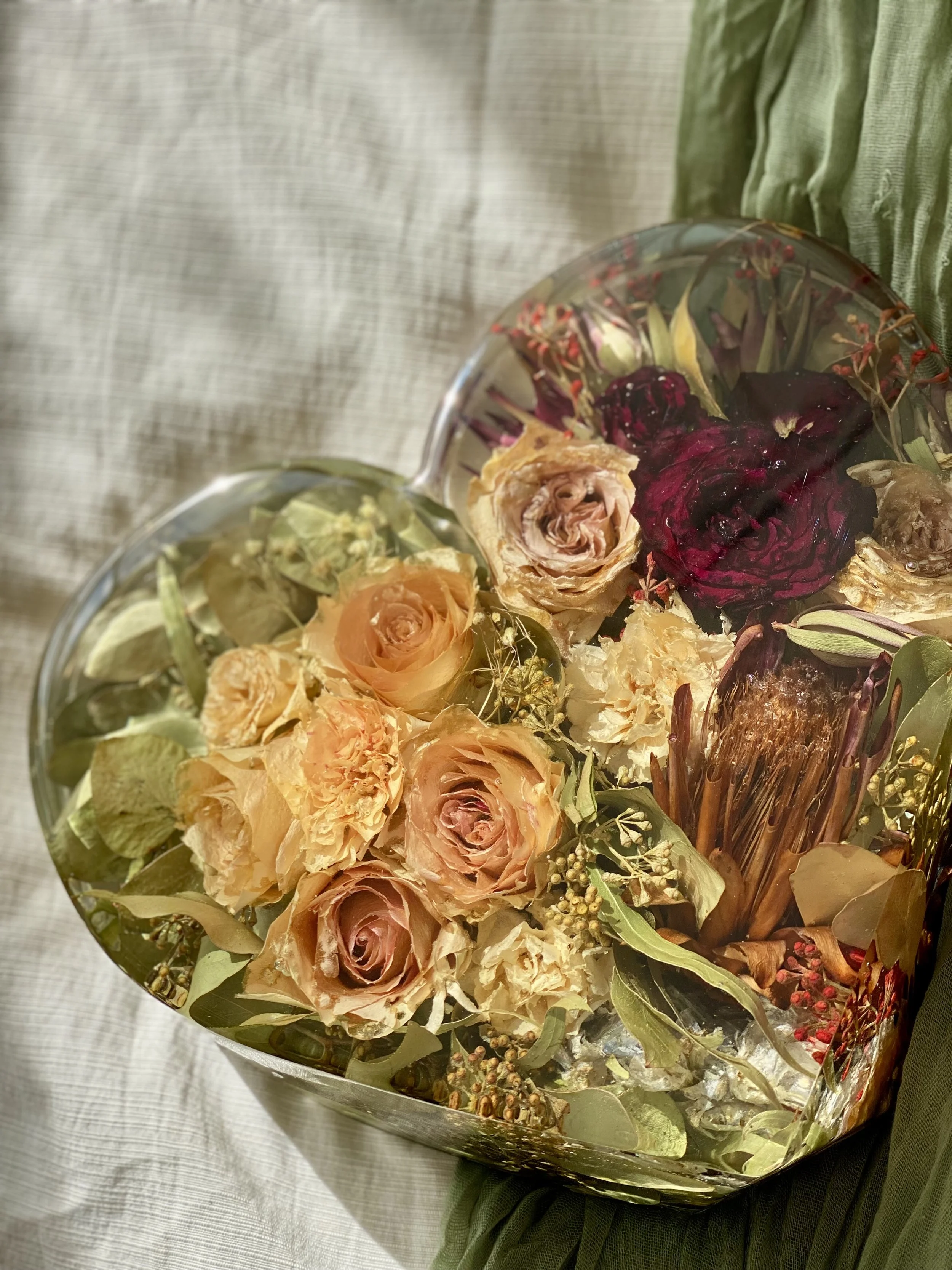 A bouquet of dried and preserved roses arranged in a silver bowl with greenery and berries, placed on a textured white surface.