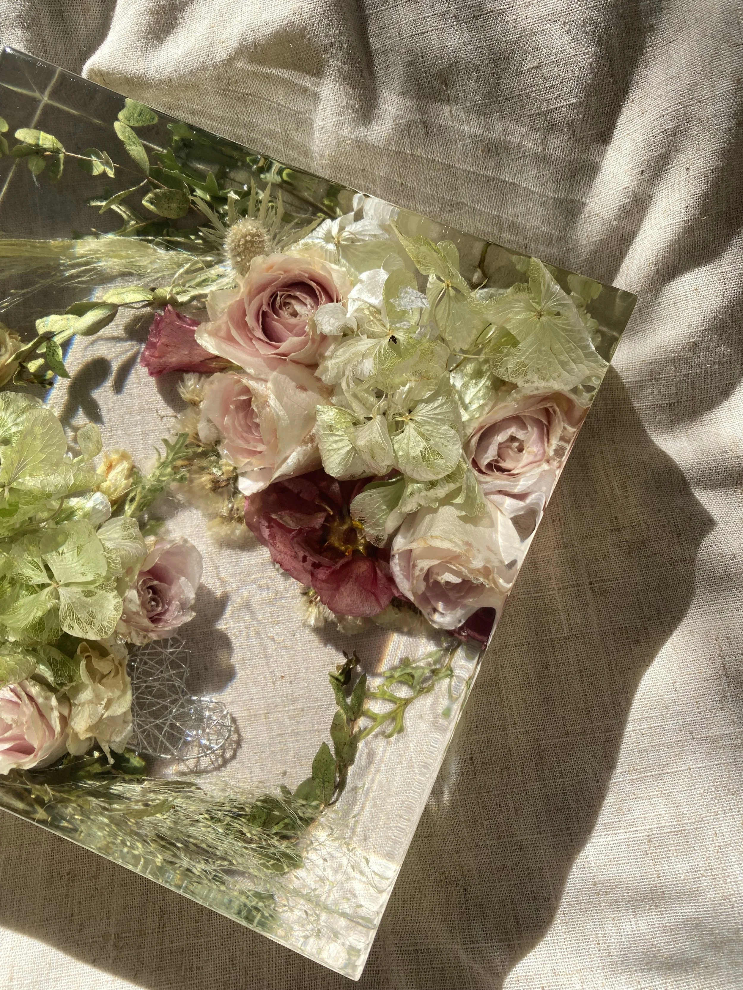 A glass tray with pink roses, white and green foliage, and decorative elements, placed on a beige fabric surface.