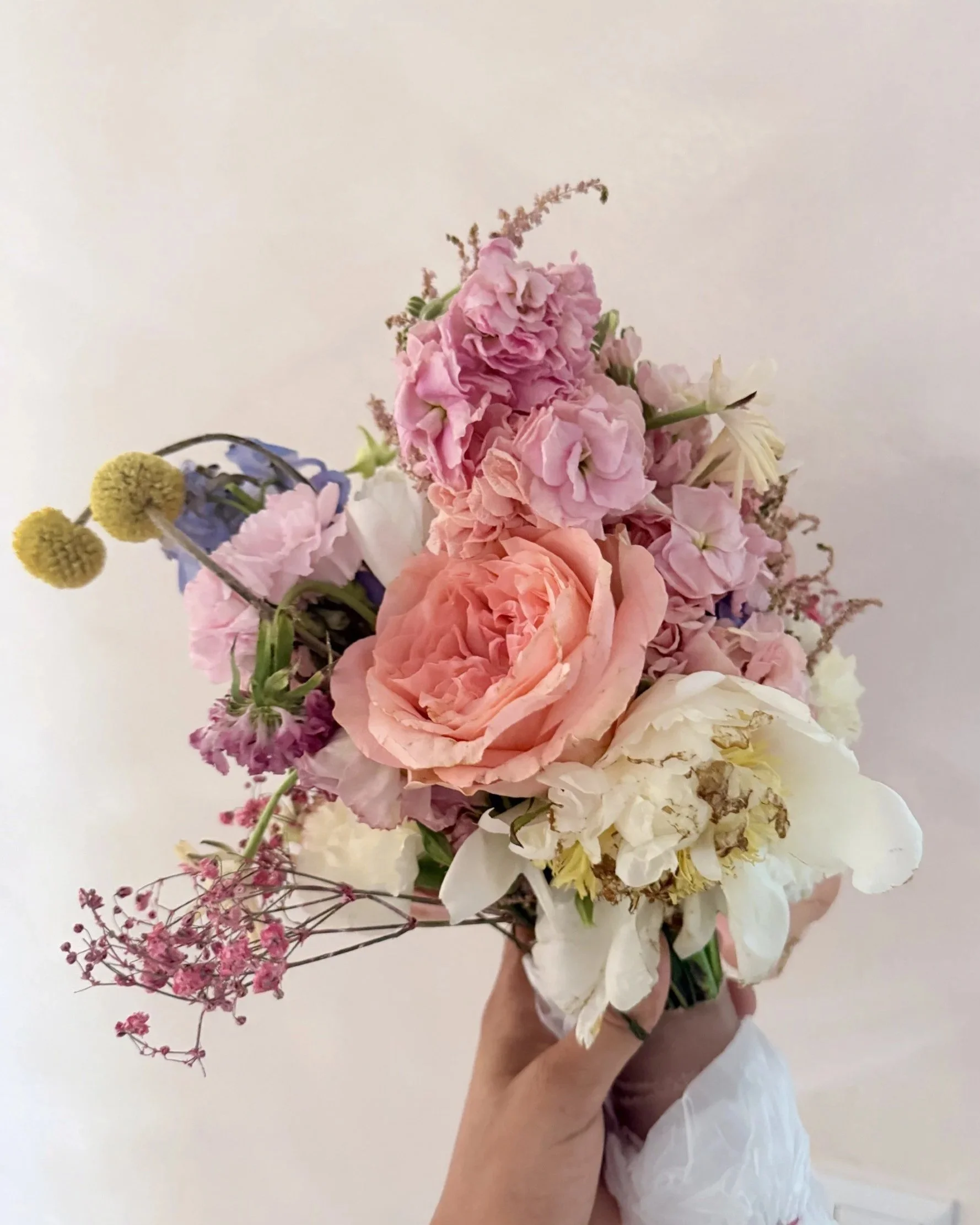A bouquet of pink, white, purple, and yellow flowers with a person holding it against a plain white background.