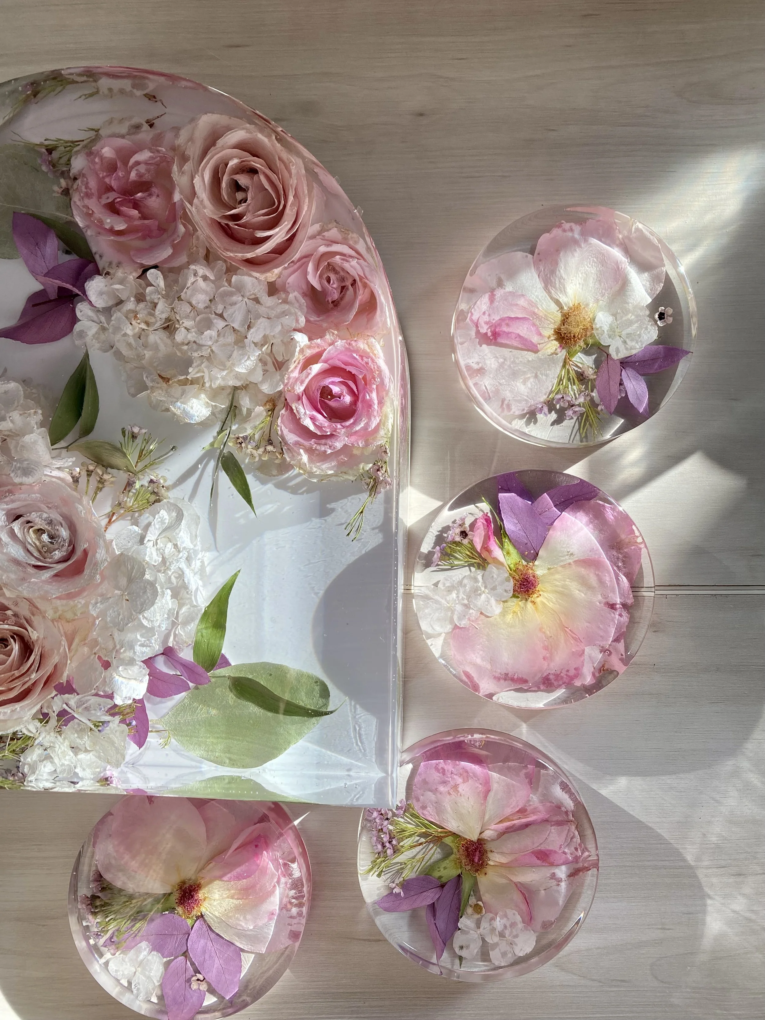 Transparent circular bowls with water containing pink roses, purple leaves, and white flowers on a wooden surface.