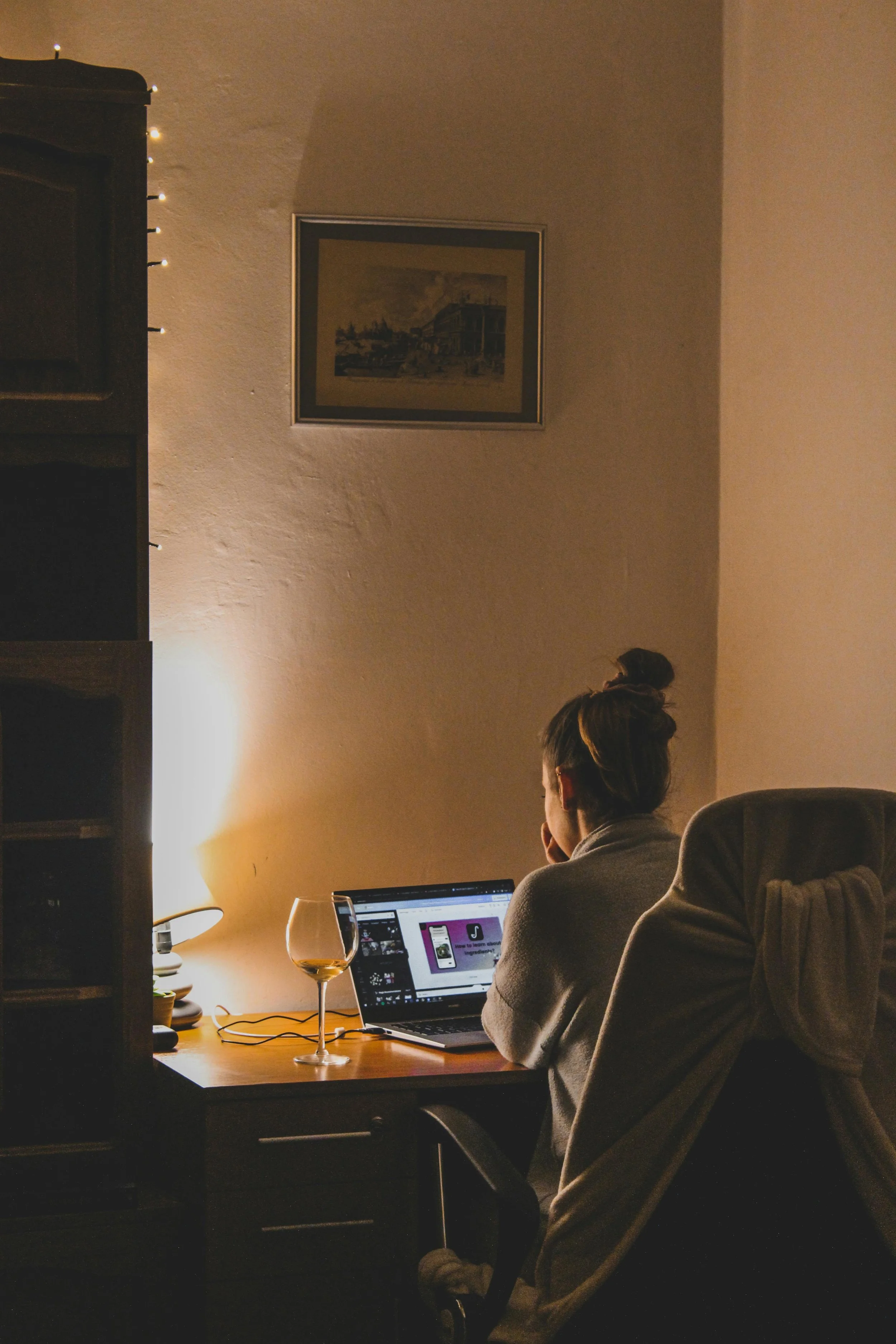 Person sitting at a desk working on a laptop in a dimly lit room, with a glass of wine and a lamp on the desk, and a framed picture on the wall.