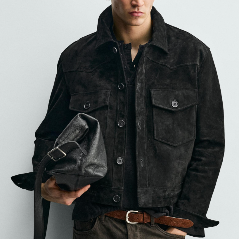 A man wearing a black denim jacket and dark pants, holding a black leather bag, standing against a plain light gray background.