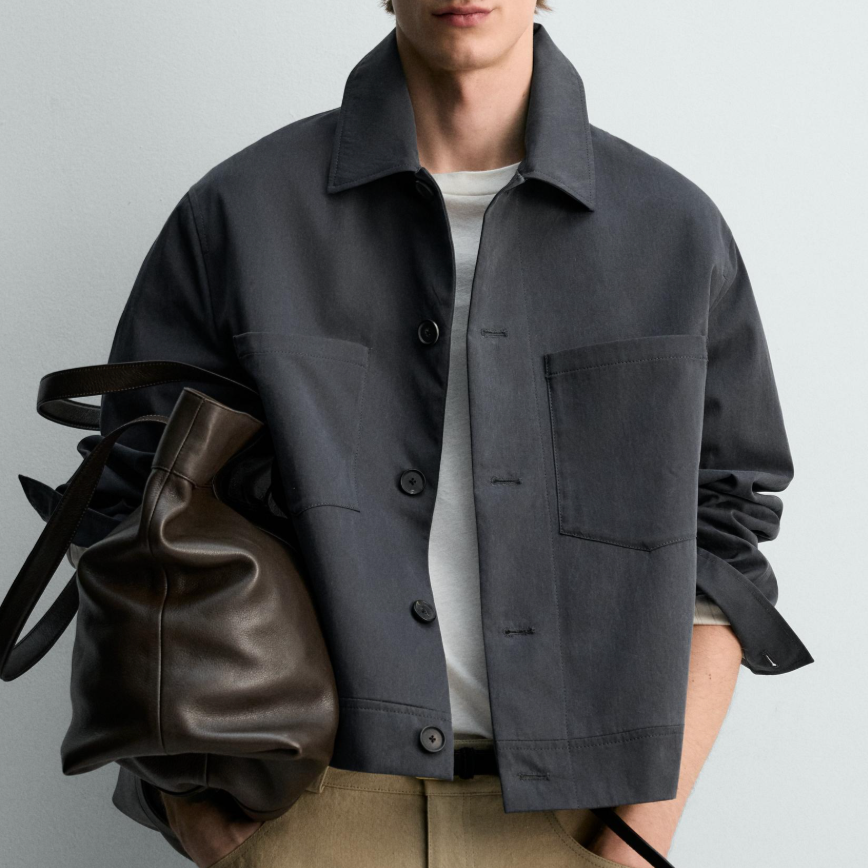 A man in a dark gray button-up jacket over a white shirt, holding a brown leather bag, standing against a light-colored wall.
