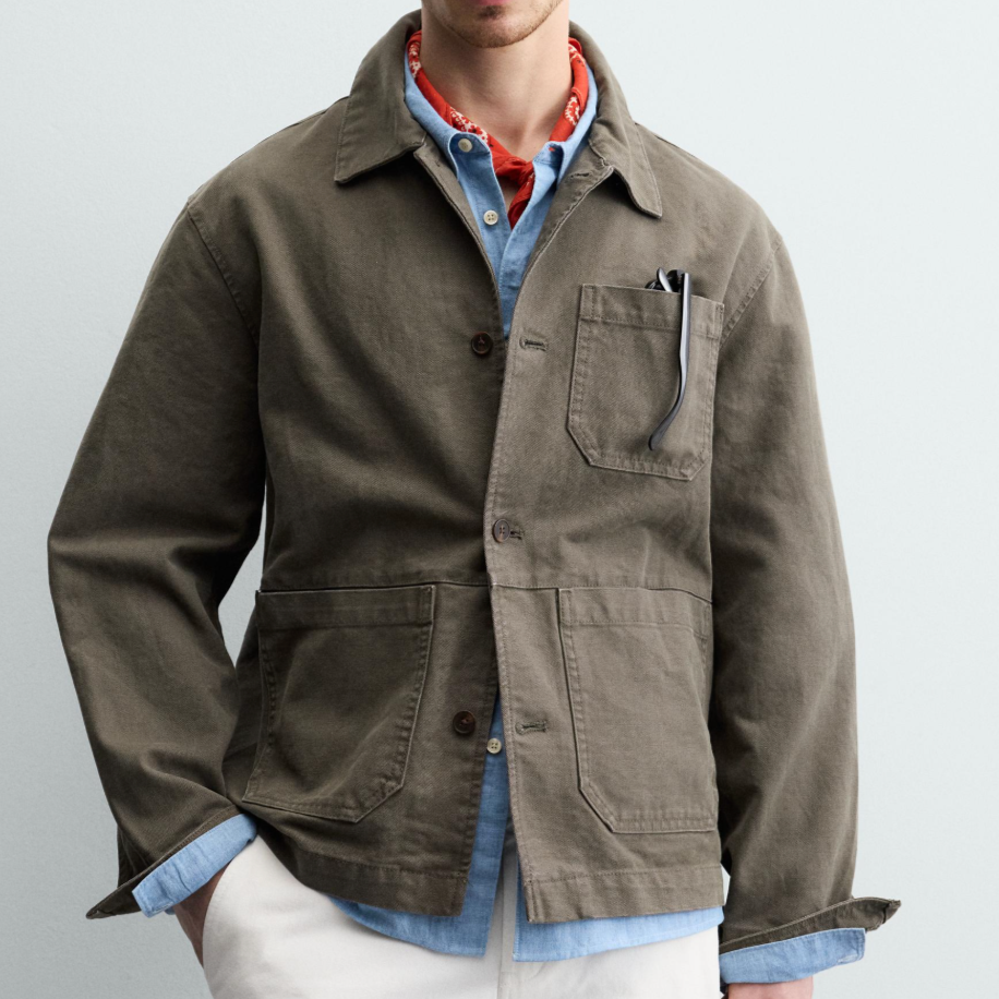 A person wearing a brown jacket with a pocket on the chest, a blue shirt underneath, and a red bandana around their neck, standing against a plain background.