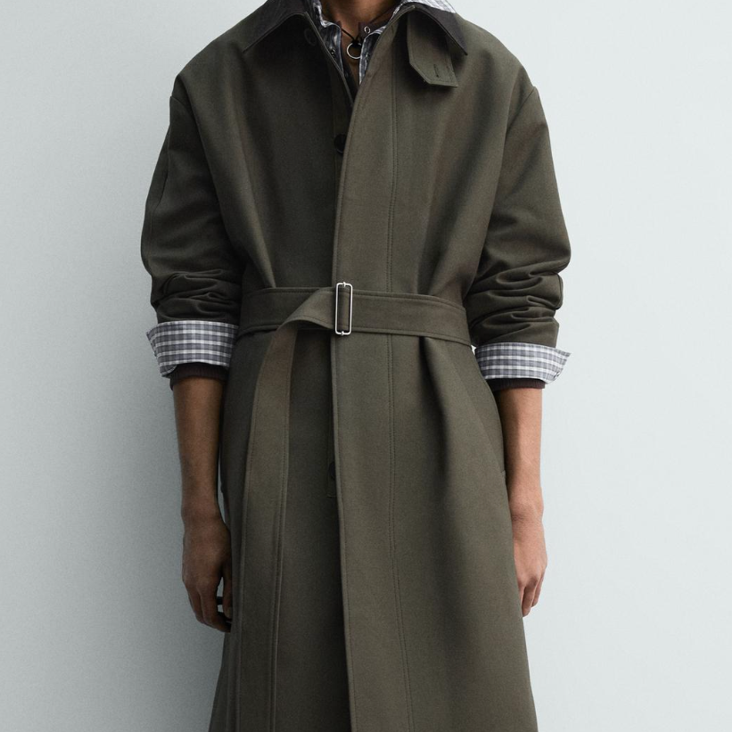 Person wearing a long, olive green trench coat with rolled-up sleeves over a checkered shirt against a plain background.