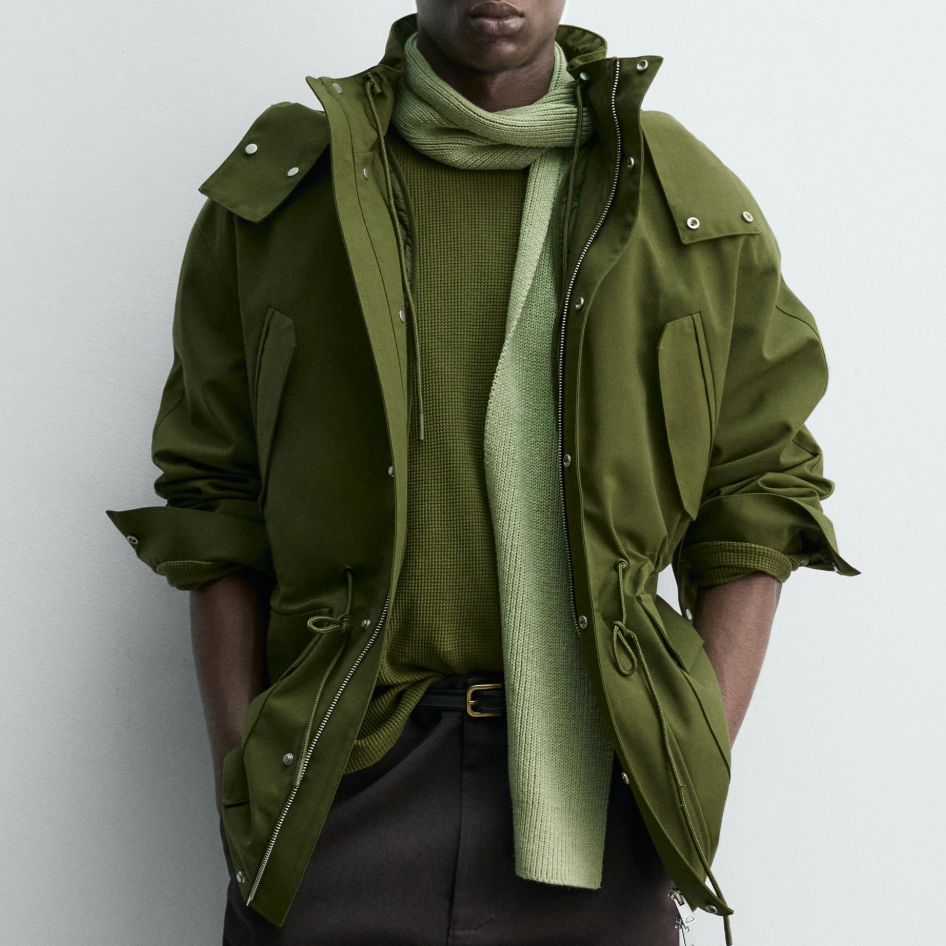 A person wearing an olive green jacket over a green textured sweater and scarf, with dark pants, standing against a gray wall.