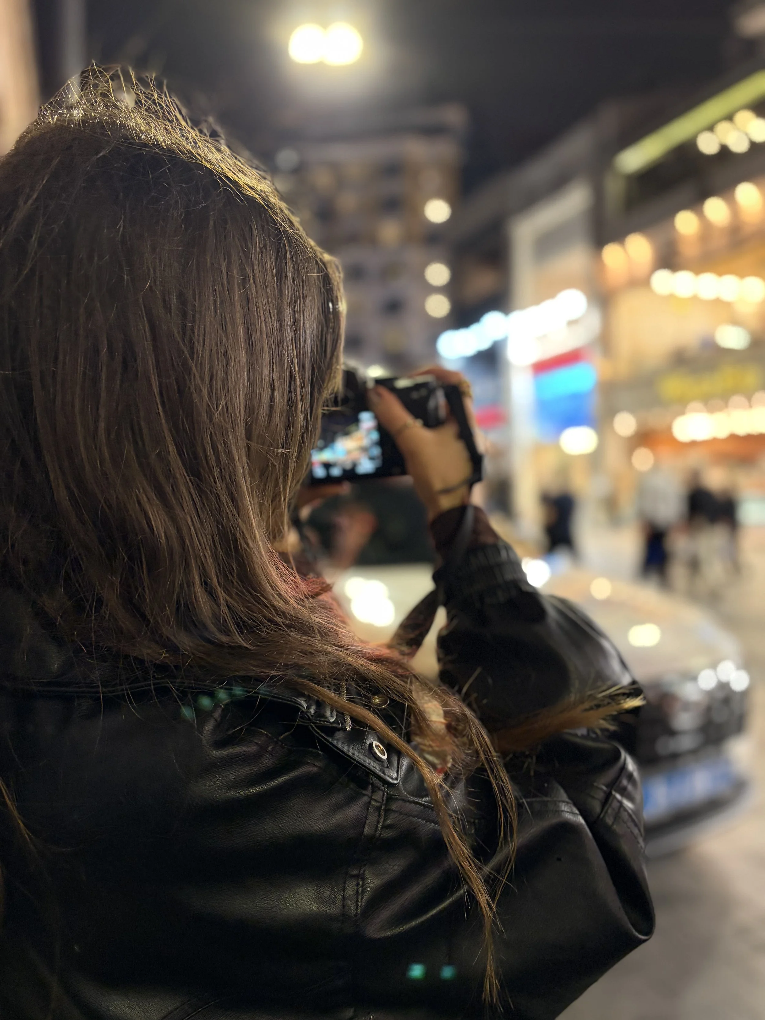 A person with long brown hair in a leather jacket taking a photo with a camera on a busy city street at night, with blurred colorful lights in the background.