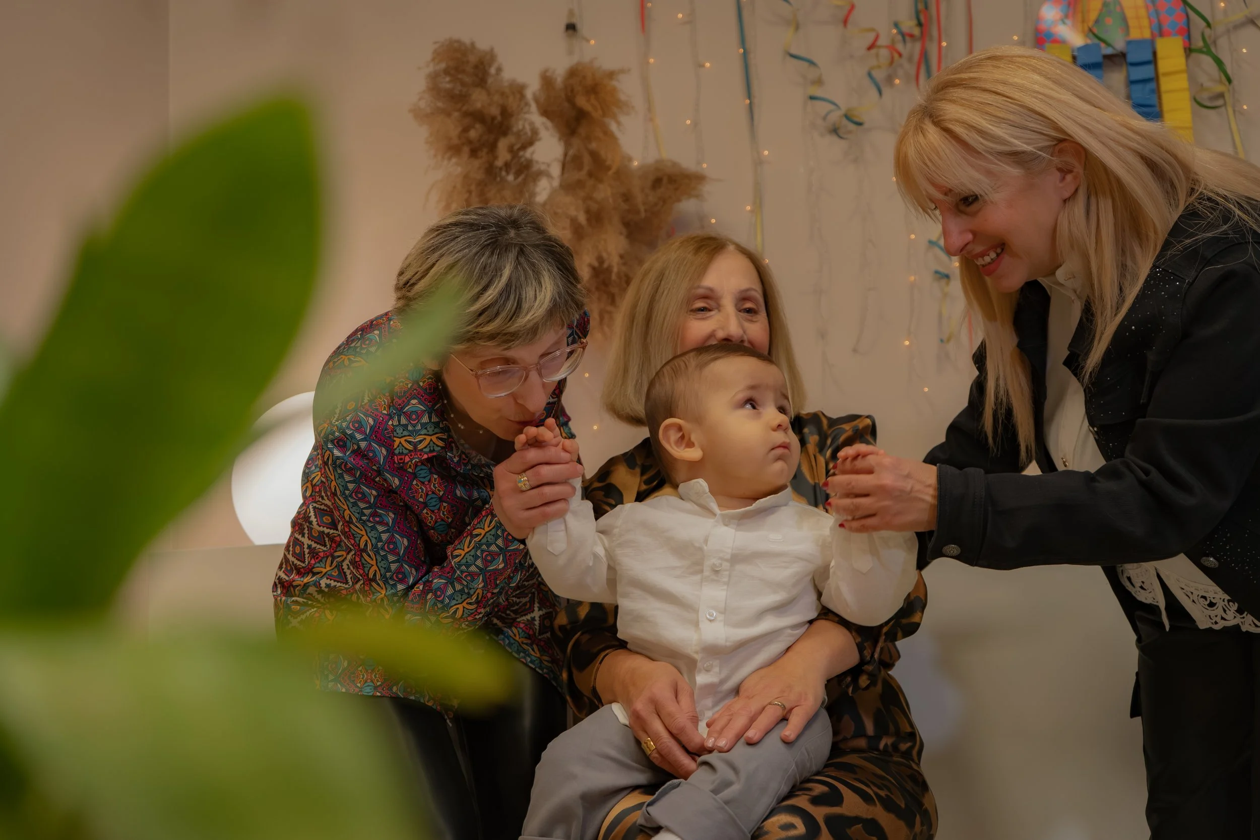A young boy in a white shirt sitting on an older woman's lap, surrounded by three women smiling and holding his hands in a festive setting with colorful decorations in the background.