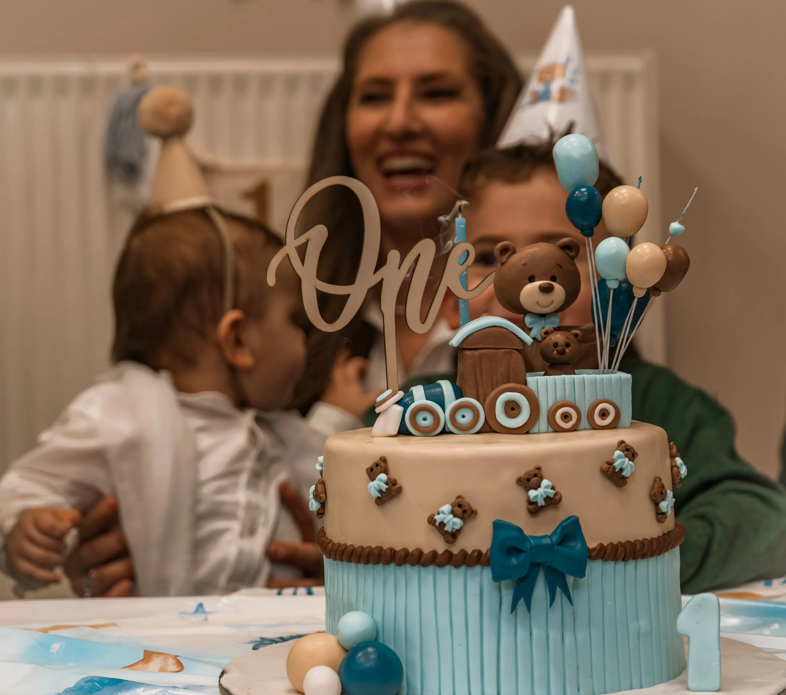 A two-tiered birthday cake with teddy bear decorations and blue, beige, and brown color scheme. A wooden train and balloons on top, with a large sign reading 'One'.