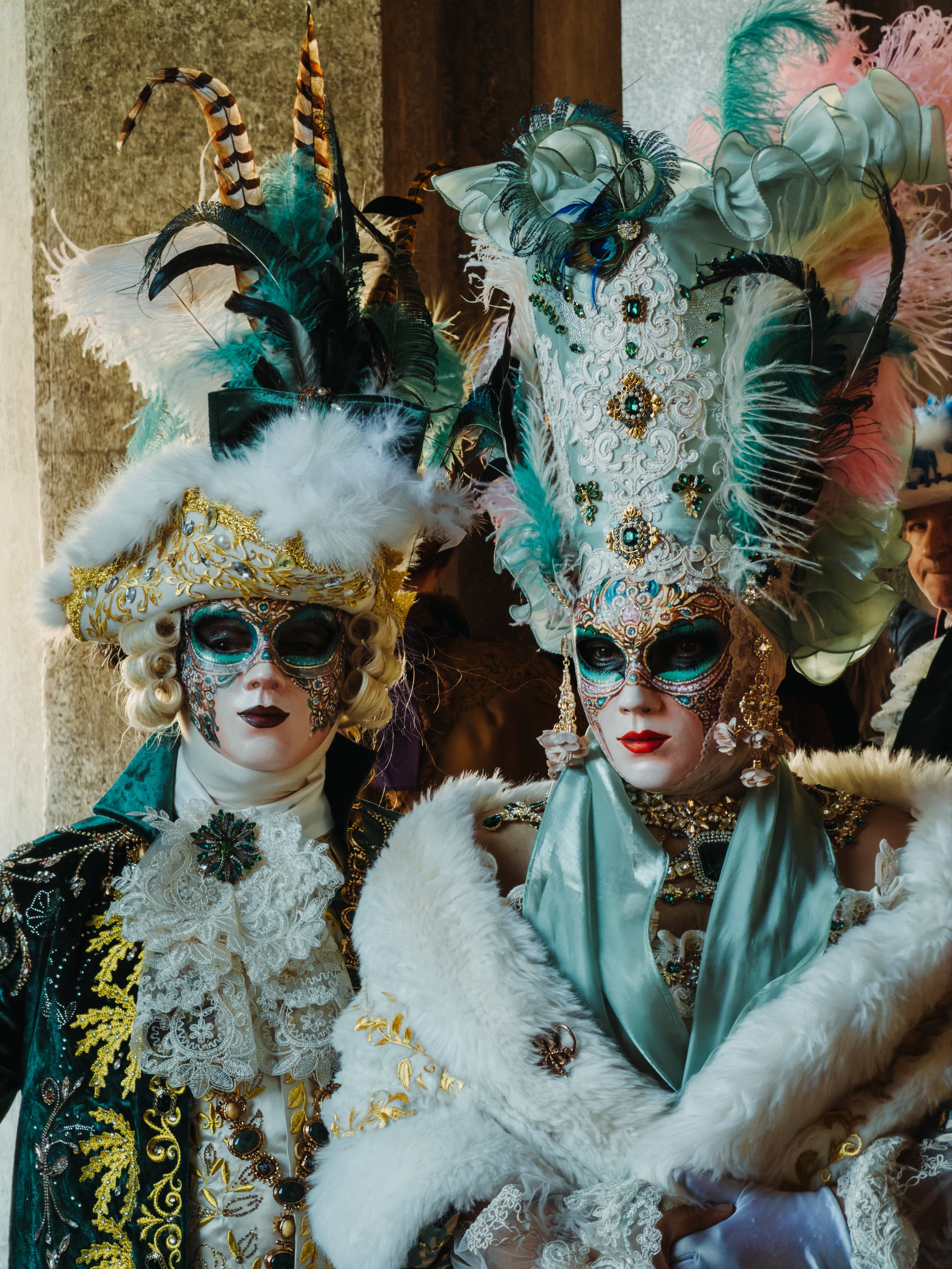 Two people dressed in elaborate, colorful costumes with ornate masks, feathers, and jewelry, likely participating in a carnival or masquerade event.