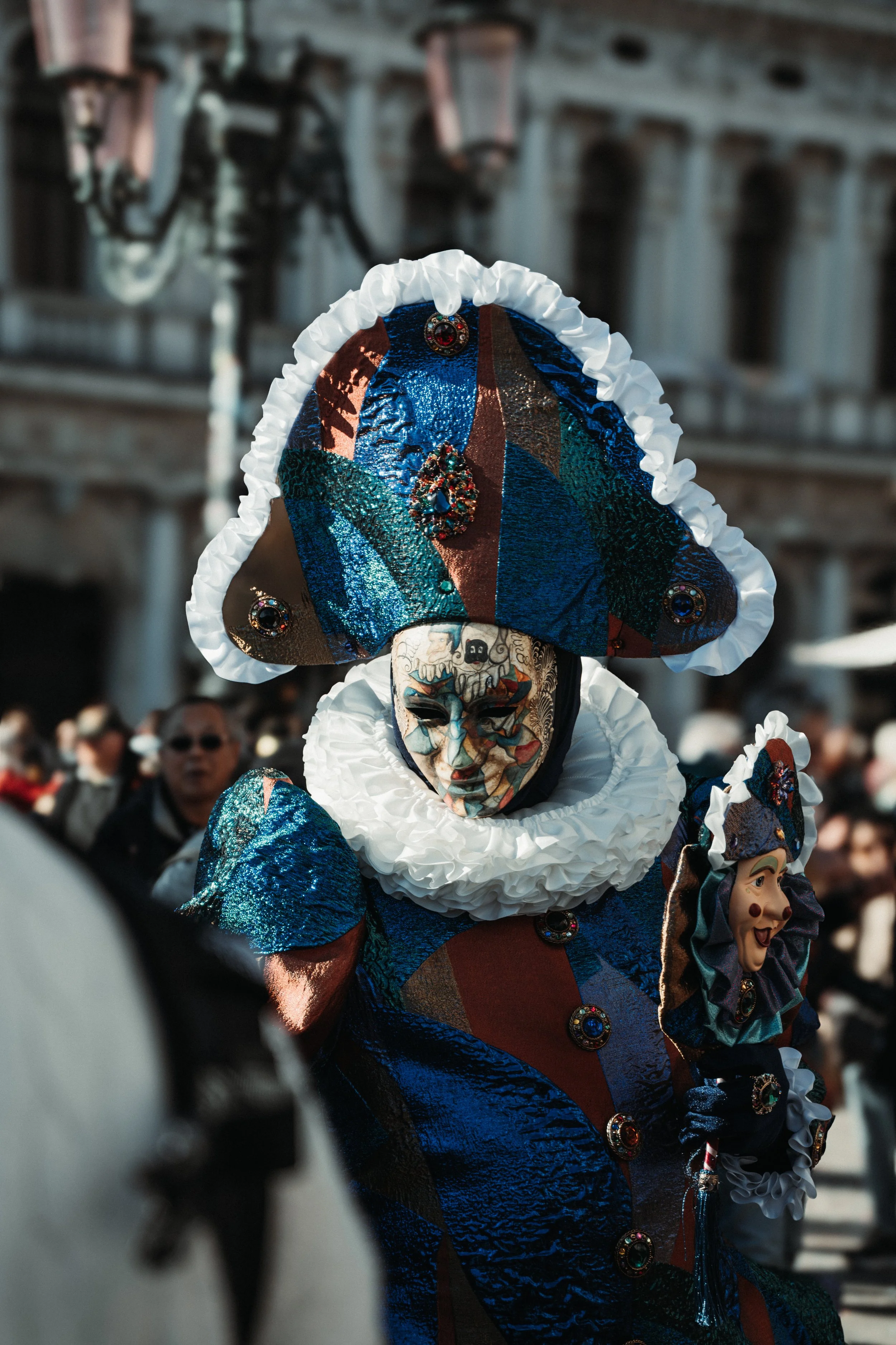 Person dressed in traditional Venetian carnival costume with a painted mask, elaborate hat, and colorful clothing, holding a puppet, in a crowded outdoor setting.