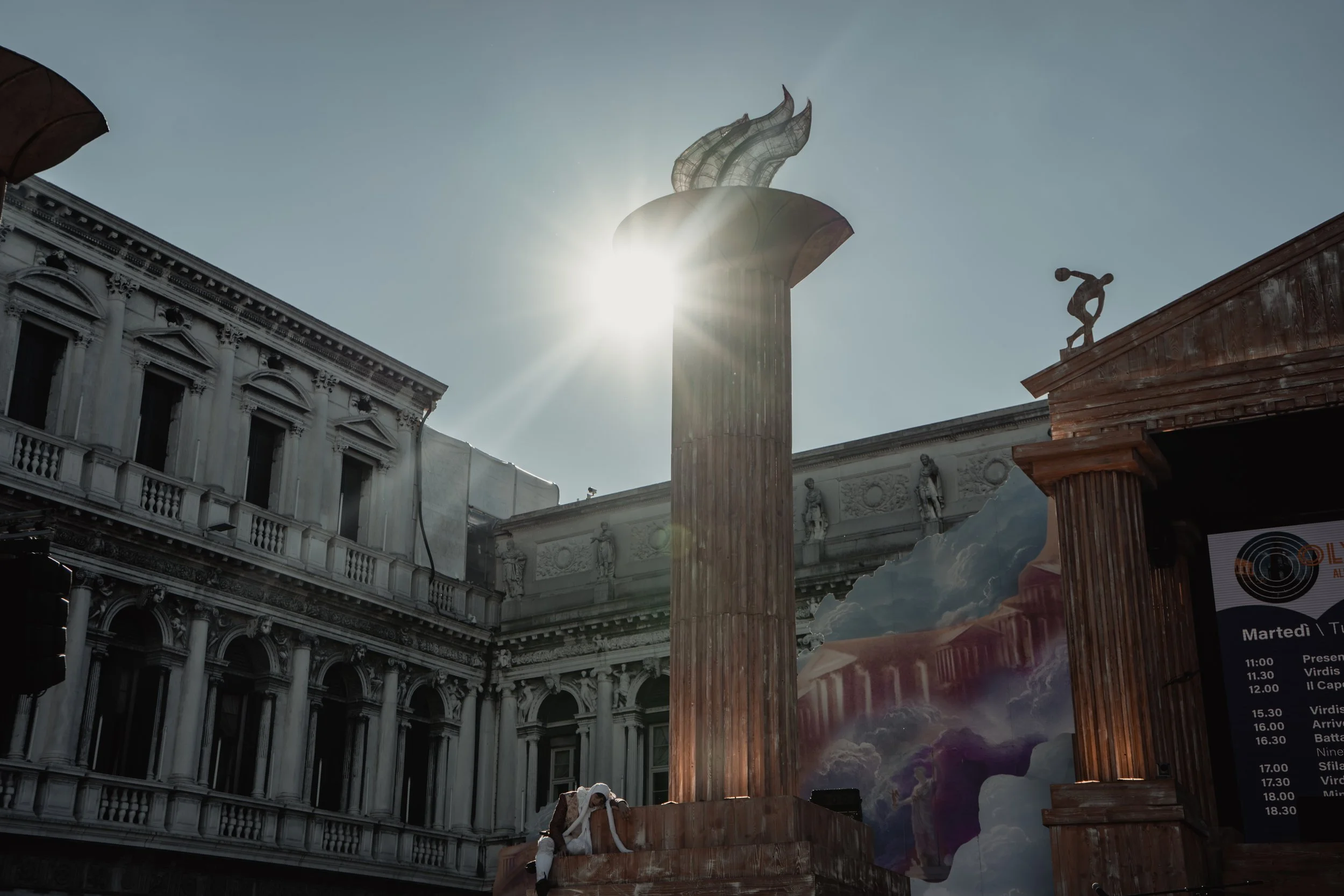 Classical architectural buildings with columns and statues, a wooden podium with a person in white leaning on it, and a backdrop with clouds and sky, with the sun shining brightly behind a column.