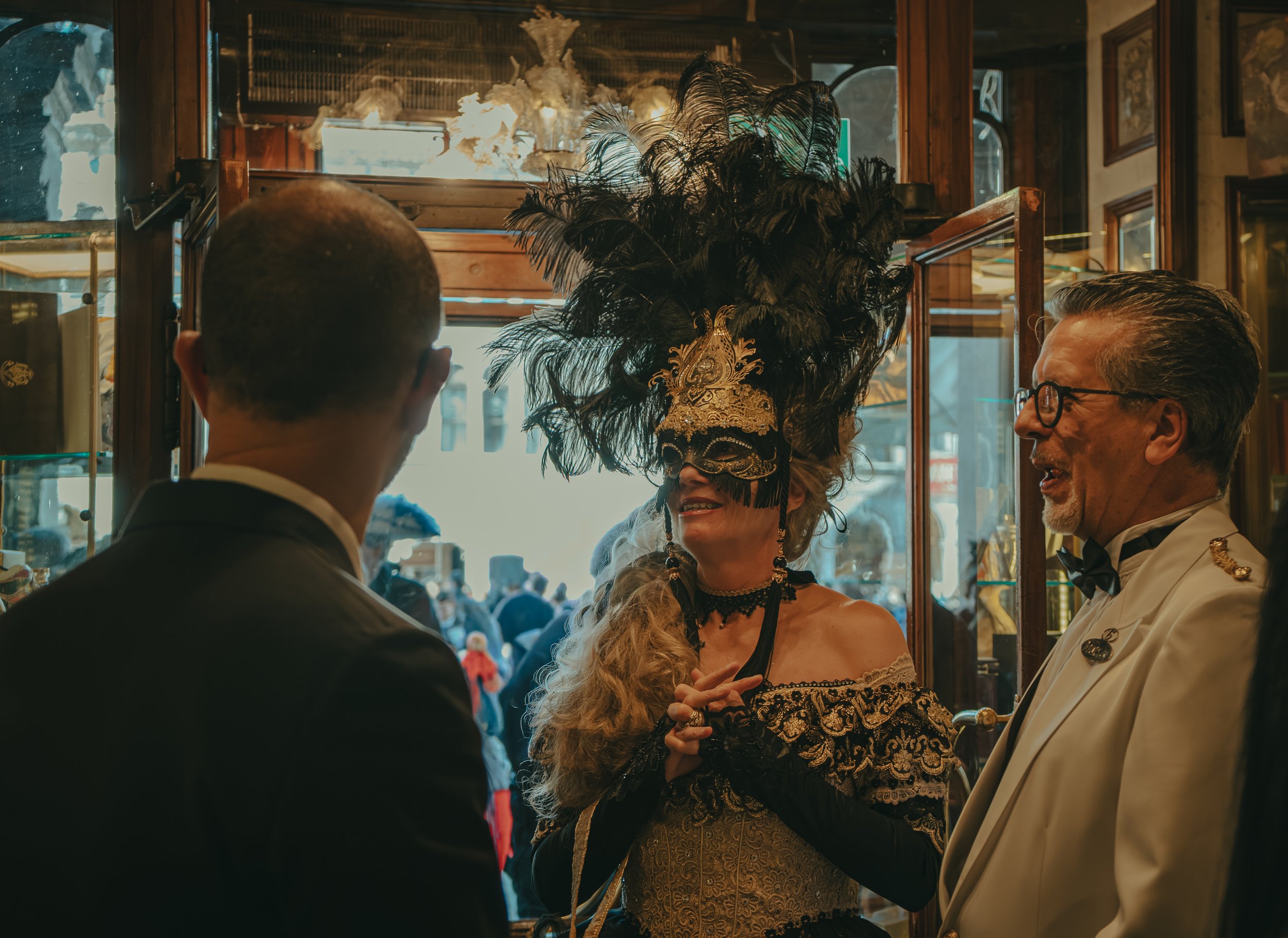 People dressed in formal attire, including a woman with an elaborate black feathered mask and gold costume, and men in tuxedos, inside a decorated room, engaging in conversation.