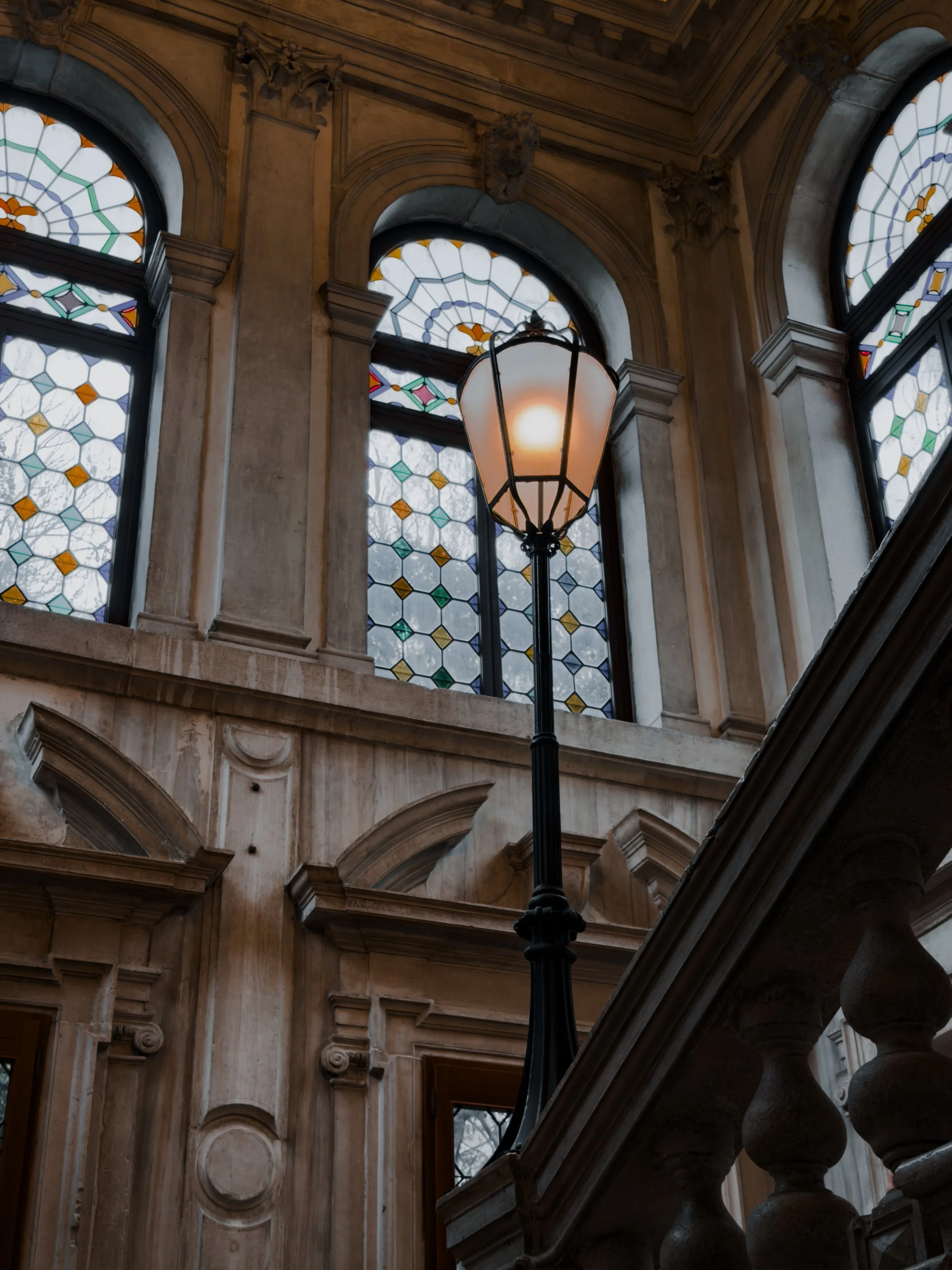 Interior view of a building with stained glass windows, a vintage lamp, and ornate architectural details.
