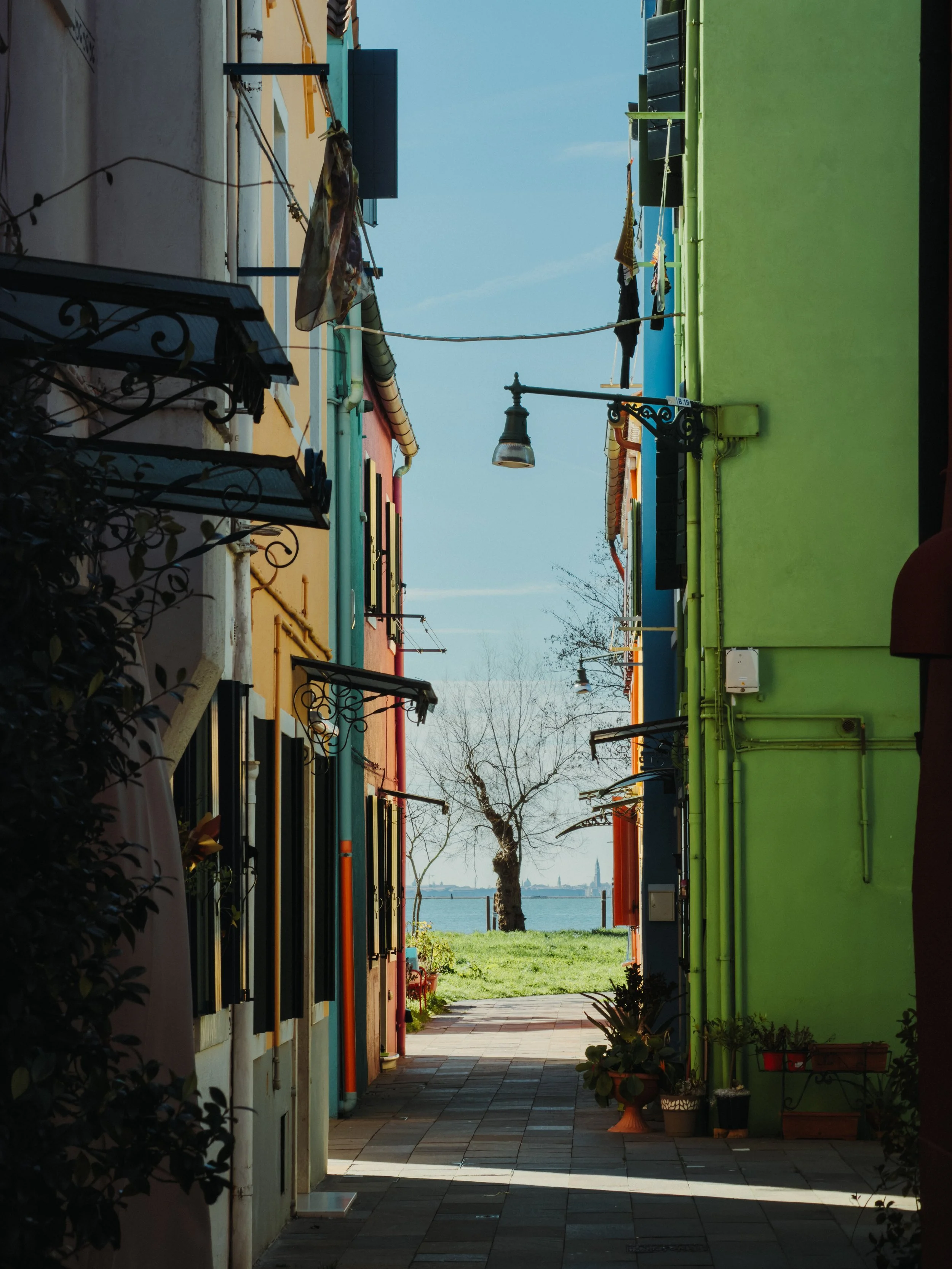 Colorful alleyway with buildings painted in pastel colors, laundry hanging to dry, and a view of water and a tree in the background.