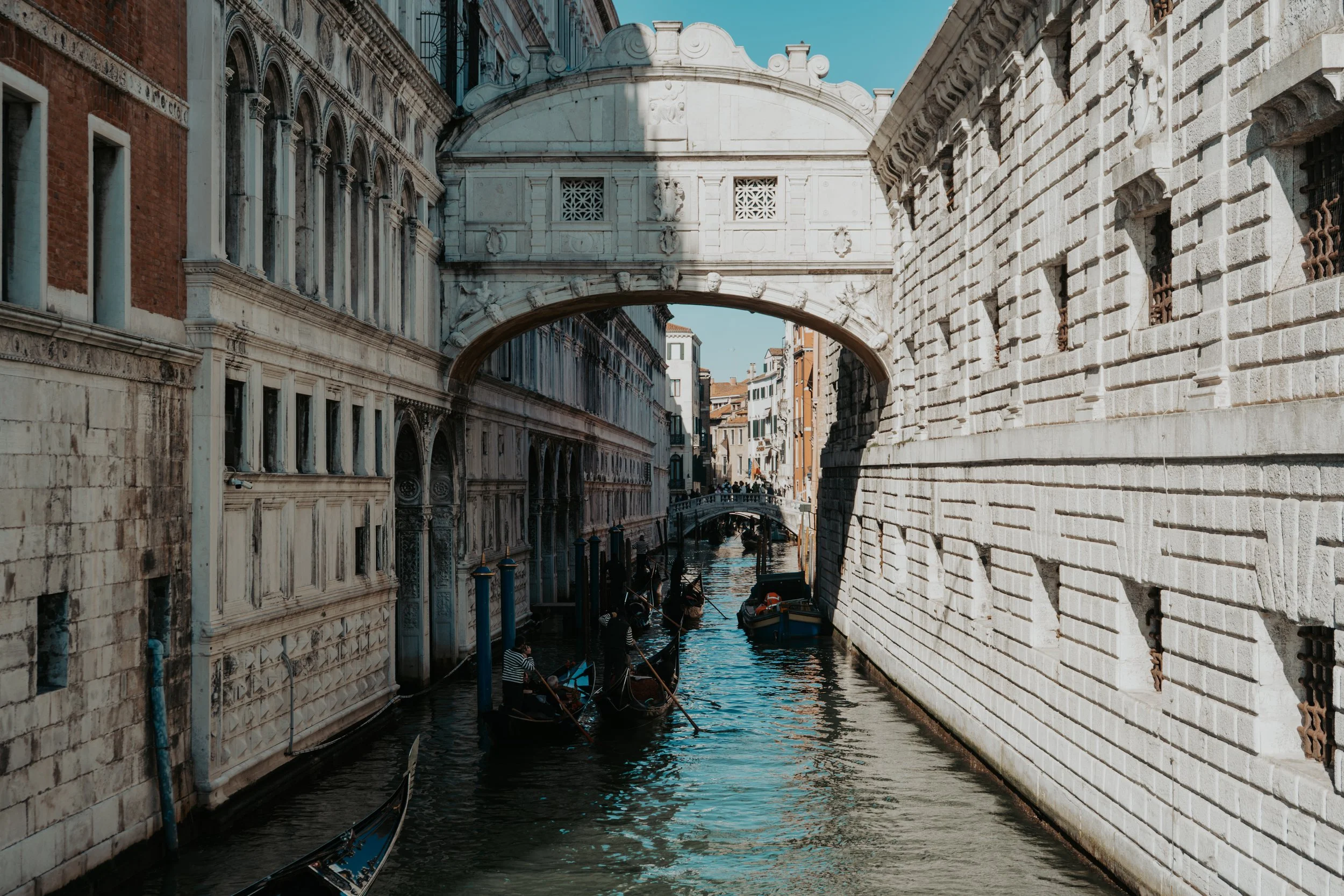 Gondolas floating on a narrow canal between historic buildings in Venice, Italy, with a small stone bridge crossing overhead.