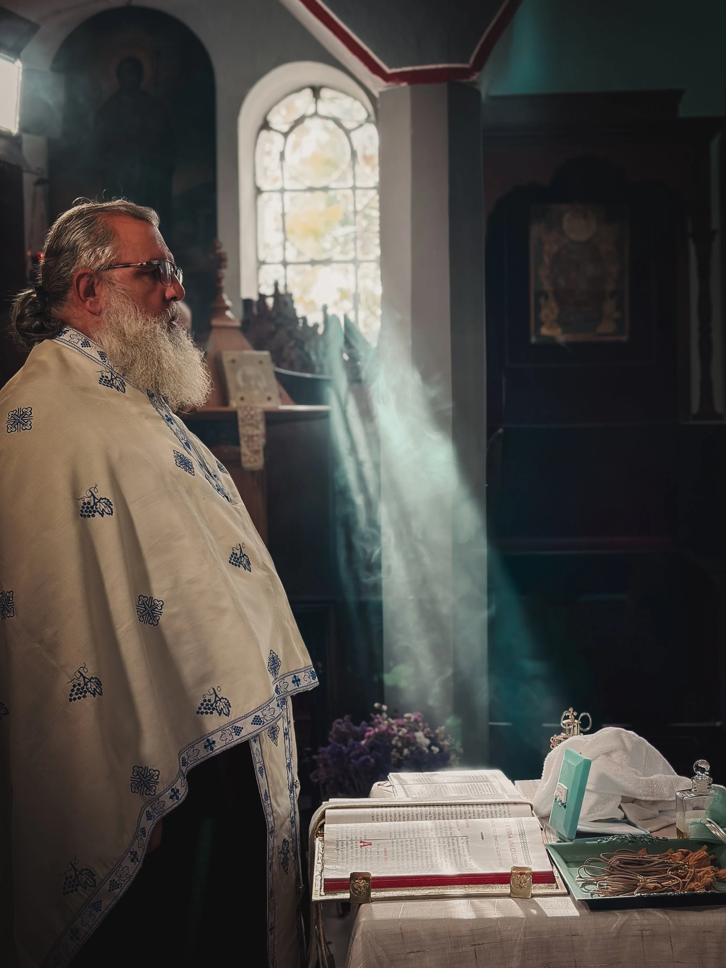 A priest with glasses and a long white beard, wearing an embroidered white vestment, standing inside a church with light streaming through a window, next to an altar with religious books and artifacts.