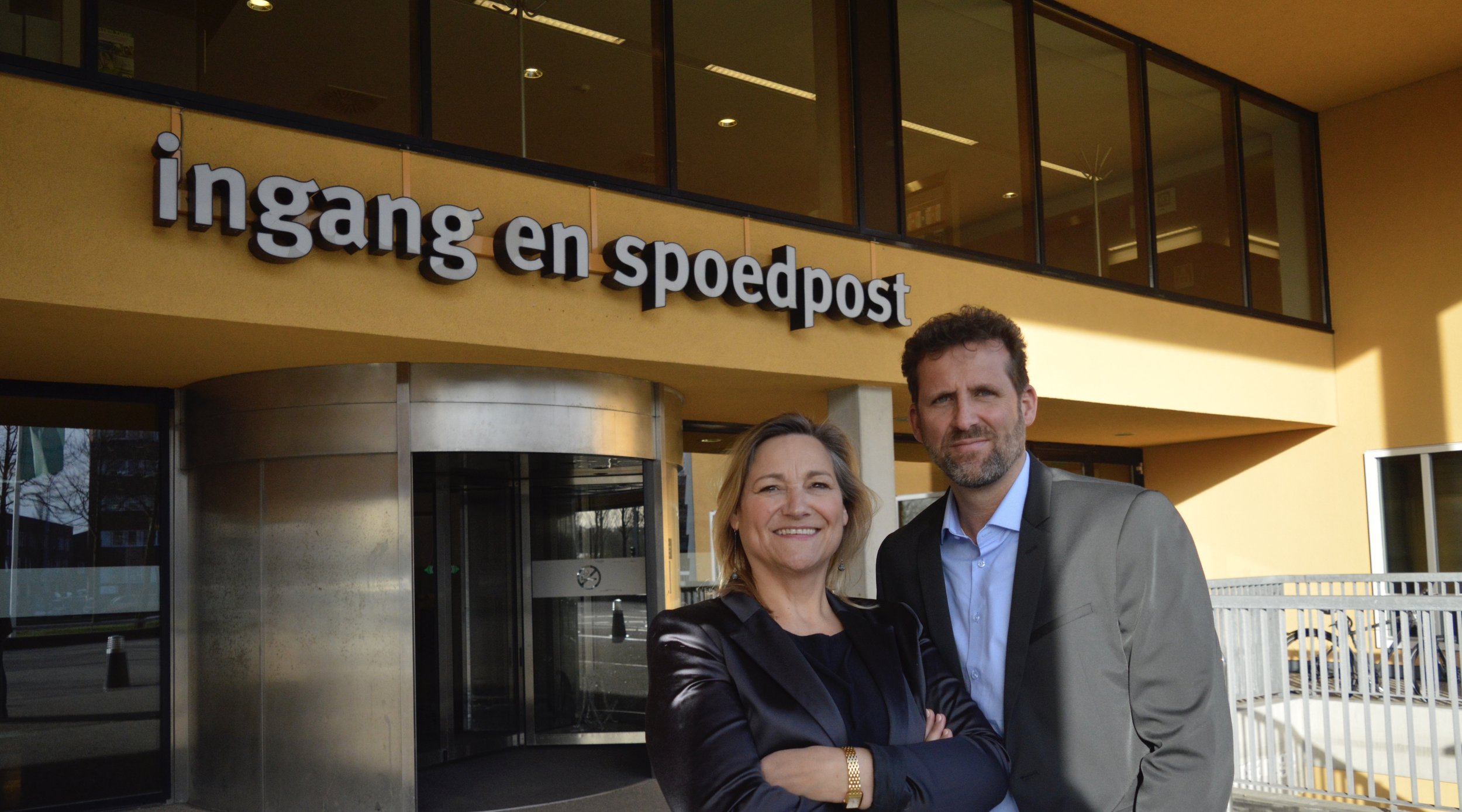 A smiling woman and man standing outside the ingang en spoedpost building, which has yellow walls and large windows.