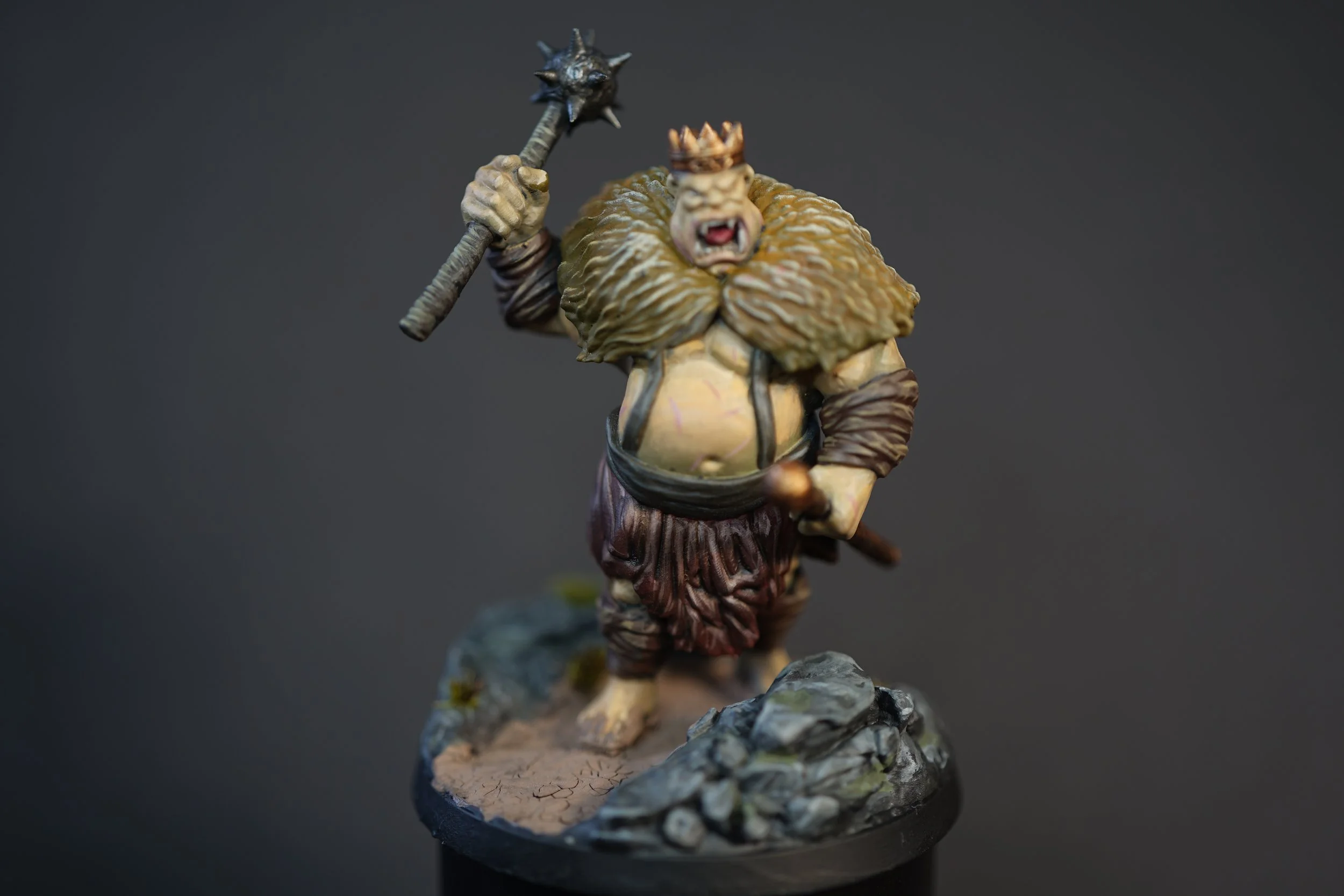 Miniature figurine of a fantasy troll with a crown, holding a spiked club, standing on a rocky base against a dark background.