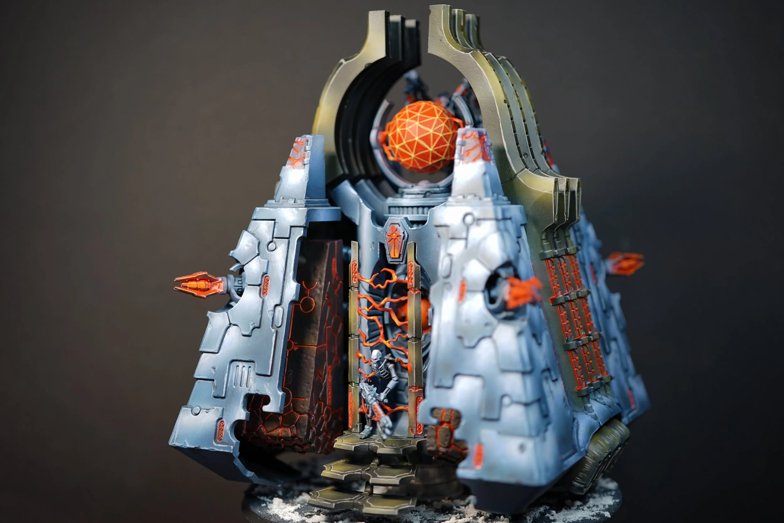 A detailed model of a futuristic, sci-fi Necron Monolith with a circular entrance, orange accents, and a floating orange and yellow geometric sphere at the top, set against a dark background.