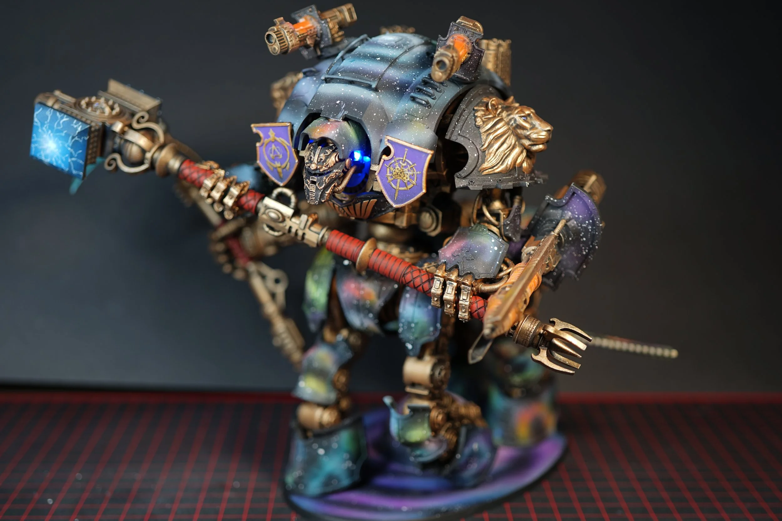 A detailed miniature model of a fantasy warrior robot, featuring armor with galaxy-like paint effects, golden lion and skull embellishments, and wielding a large warhammer, axe and spear.