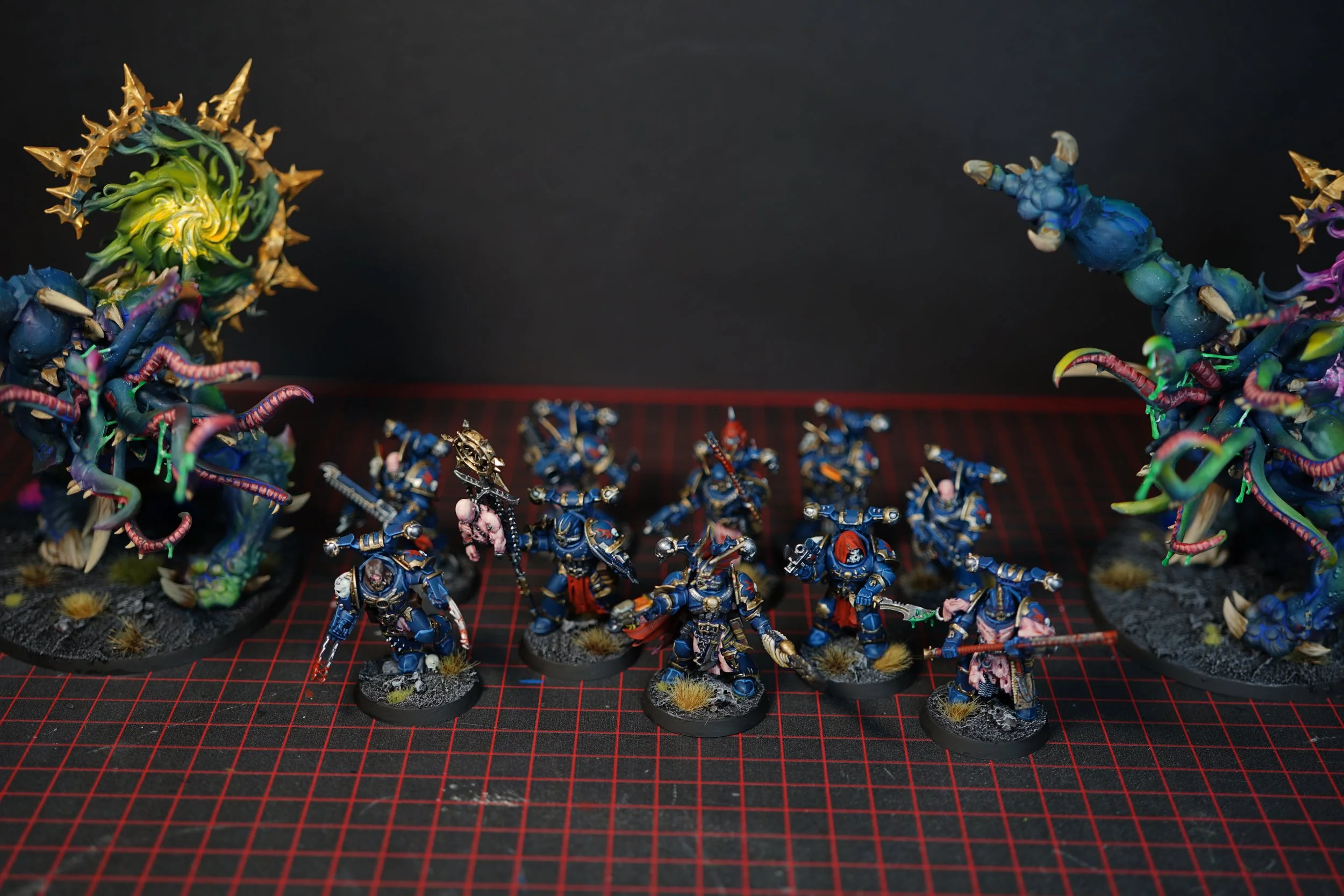 Group of tabletop miniature soldiers in blue armor with weapons, surrounded by large, colorful, tentacle-like creatures on a red grid cutting mat.