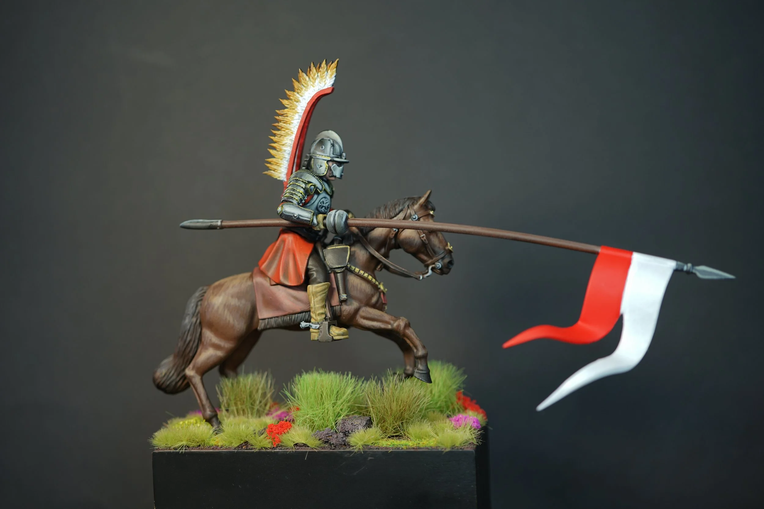 A detailed miniature figurine of a medieval polish hussar on horseback, equipped with armor, a helmet, a colorful plume, and carrying a lance with a red and white banner, set on a grassy base.