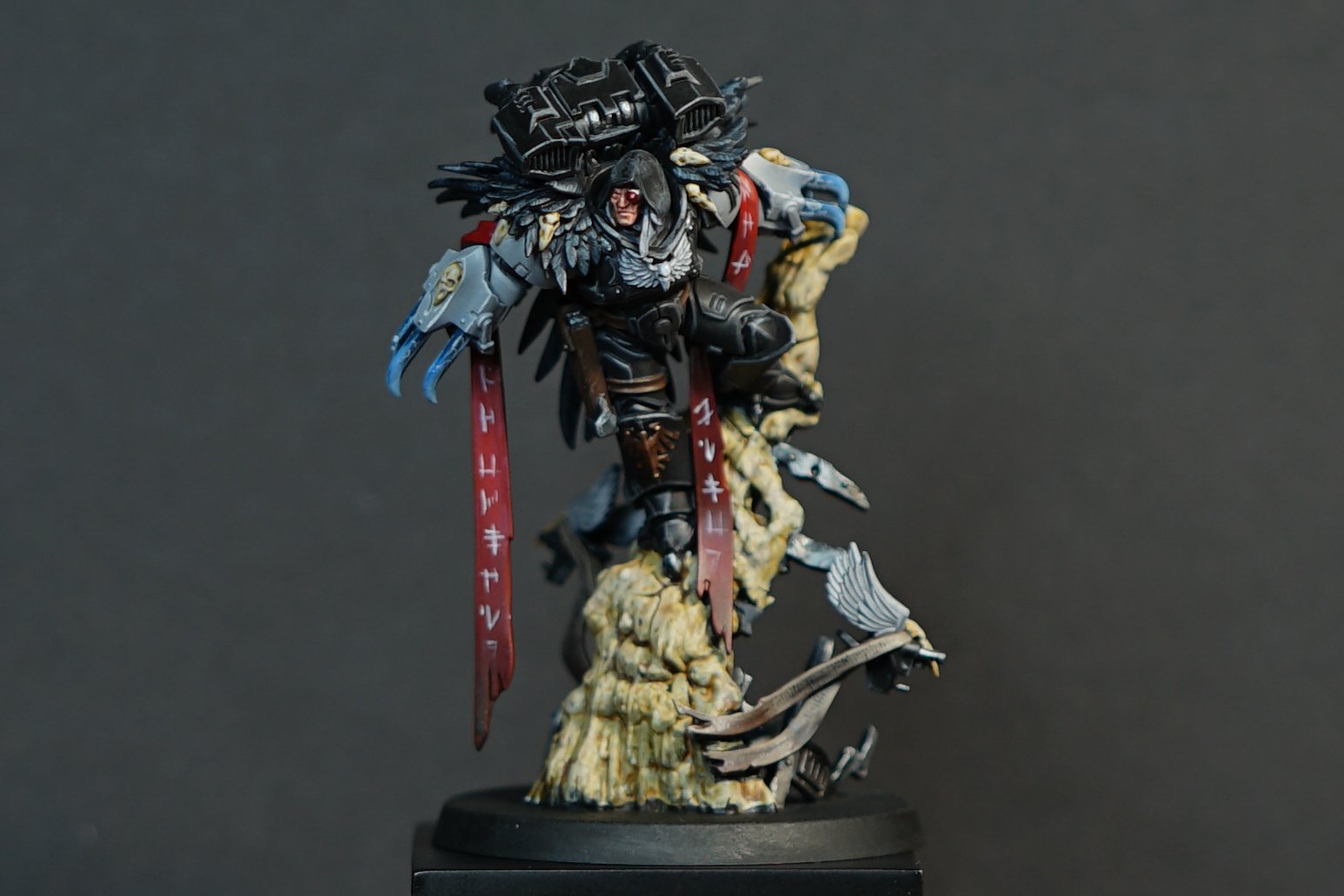 Close-up of a detailed painted miniature figurine of a Raven Guard captain from Warhammer 40k with feathers, wielding two energy claws, standing on a terrain base.