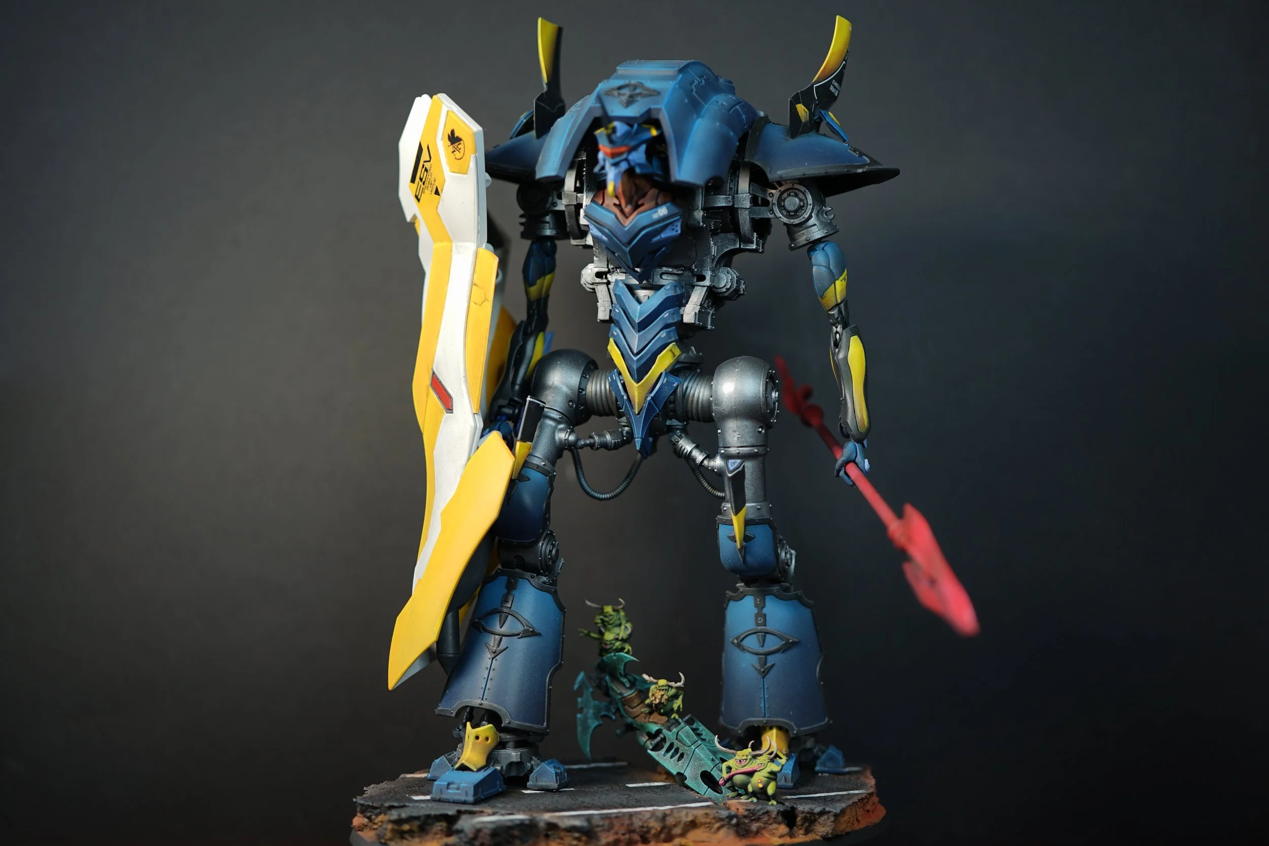 A detailed model of a large robot with blue, yellow, and black armor, holding a shield and a spear, standing on a textured base with small green creatures.