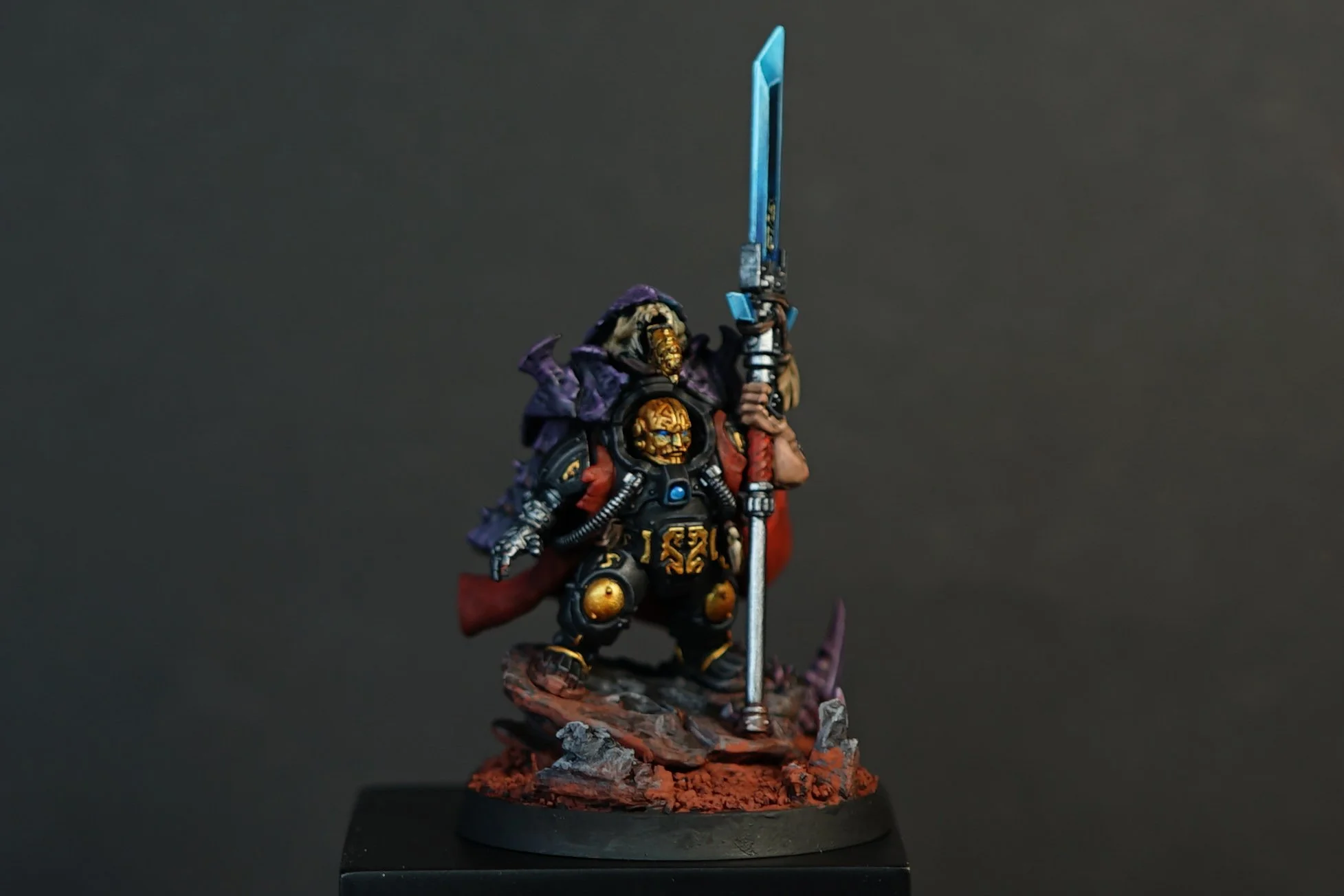A painted miniature of Buri Aegrnisson from the Leagues of Votann in Warhammer 40k with a golden face mask, dressed in black armor with gold accents, wearing a red cloak and holding a large blue spear, standing on a textured base with rocks and dirt.