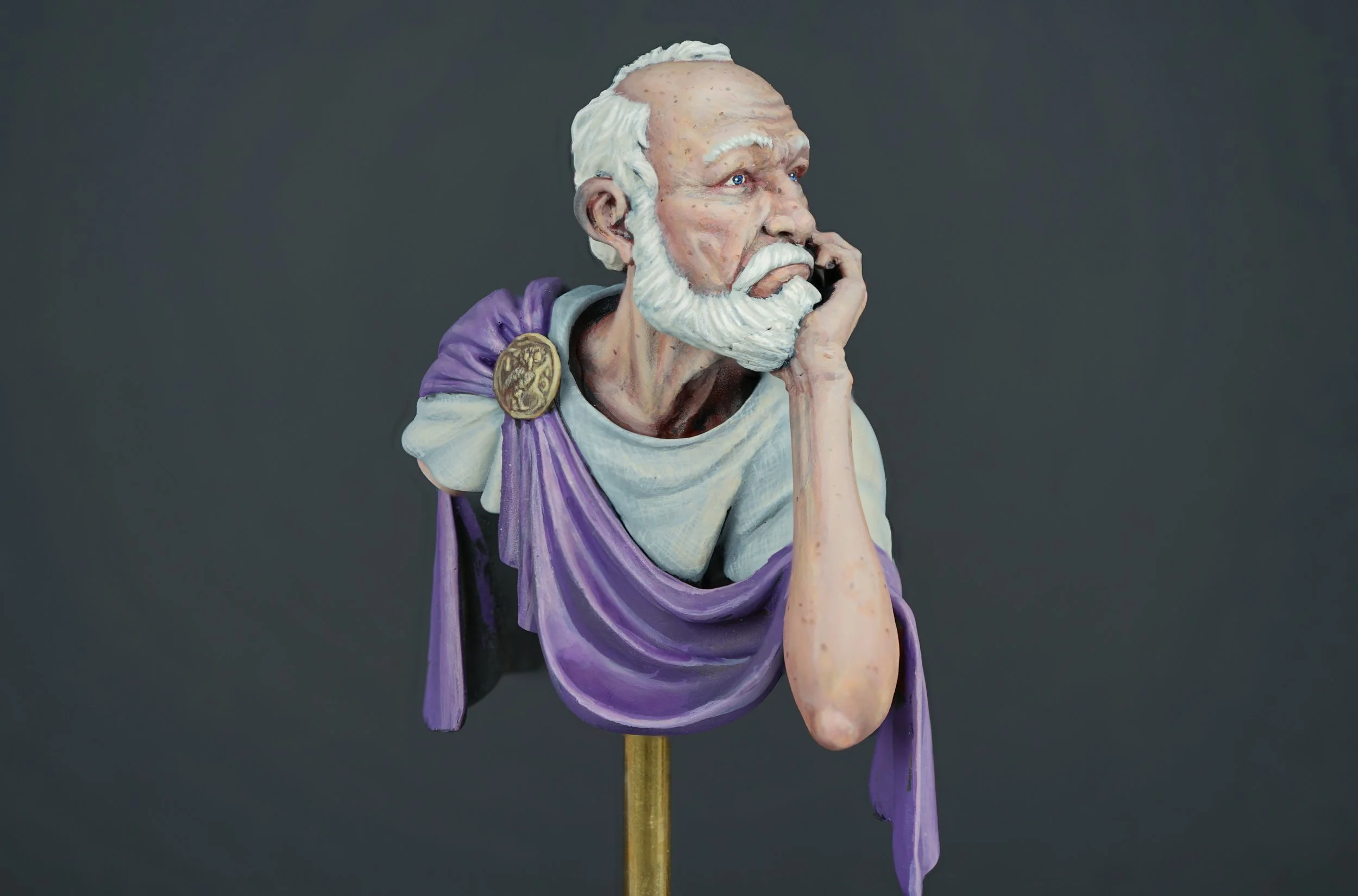 A realistic sculpture of an elderly man with white hair and beard, wearing a purple toga with a gold brooch, resting his chin on his hand and appearing to be deep in thought.