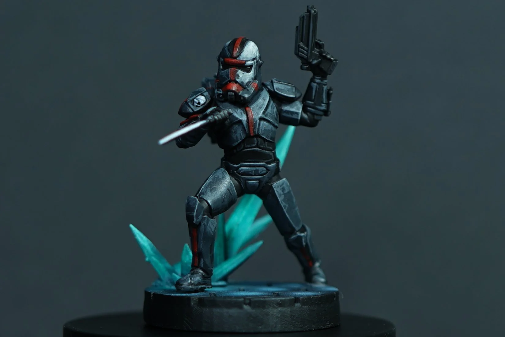 A detailed Star Wars Legion miniature figurine of Hunter from the clone trooper unit Bad Batch in black armor with red and silver accents, holding a gun in one hand and a blade in the other, standing on a base with blue crystal-like formations.