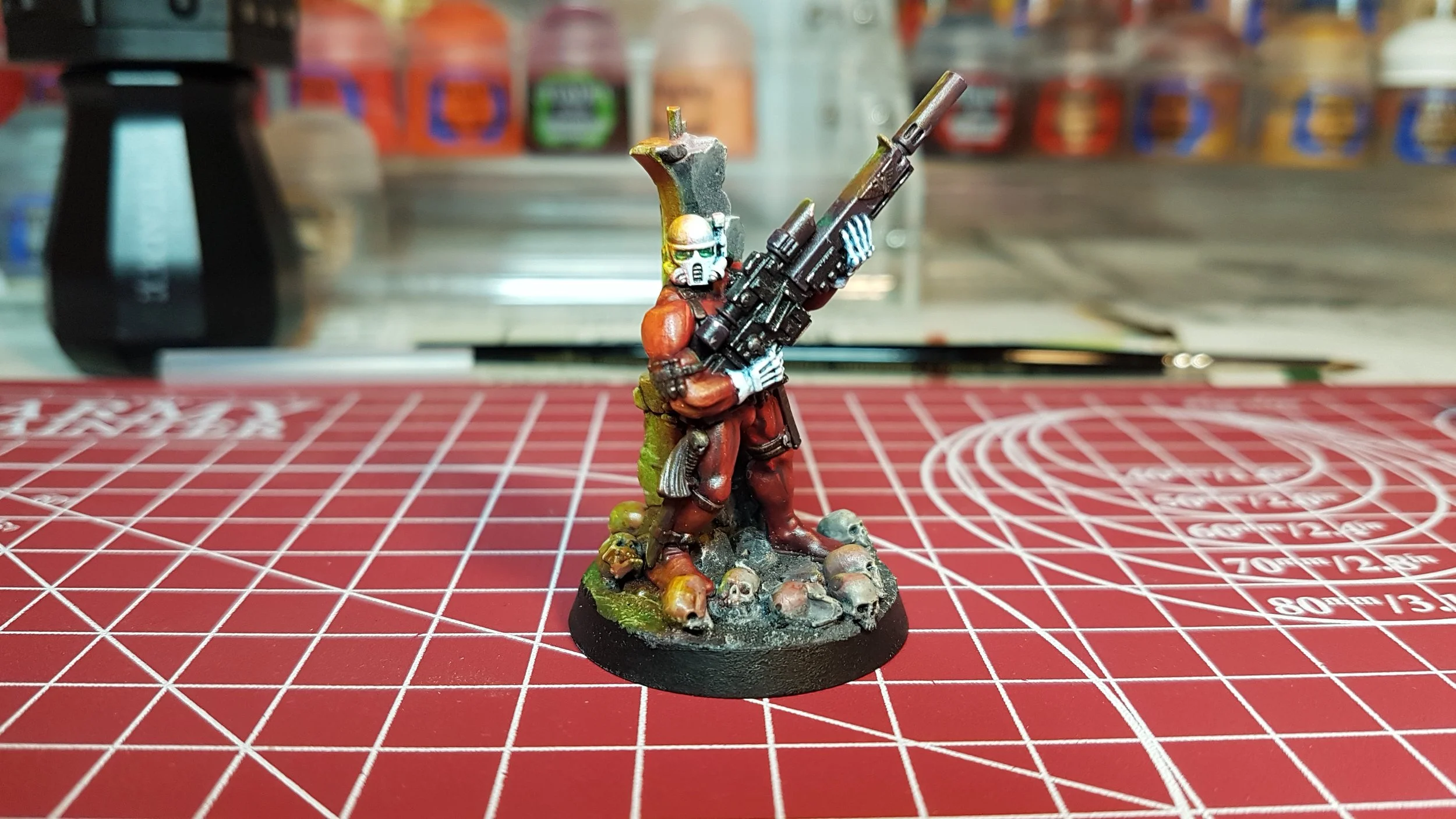 A painted miniature figurine of a sci-fi soldier in red armor with a skull helmet, holding a large rifle, standing on a war-torn base with skulls and debris, on a red cutting mat with measuring grid lines.