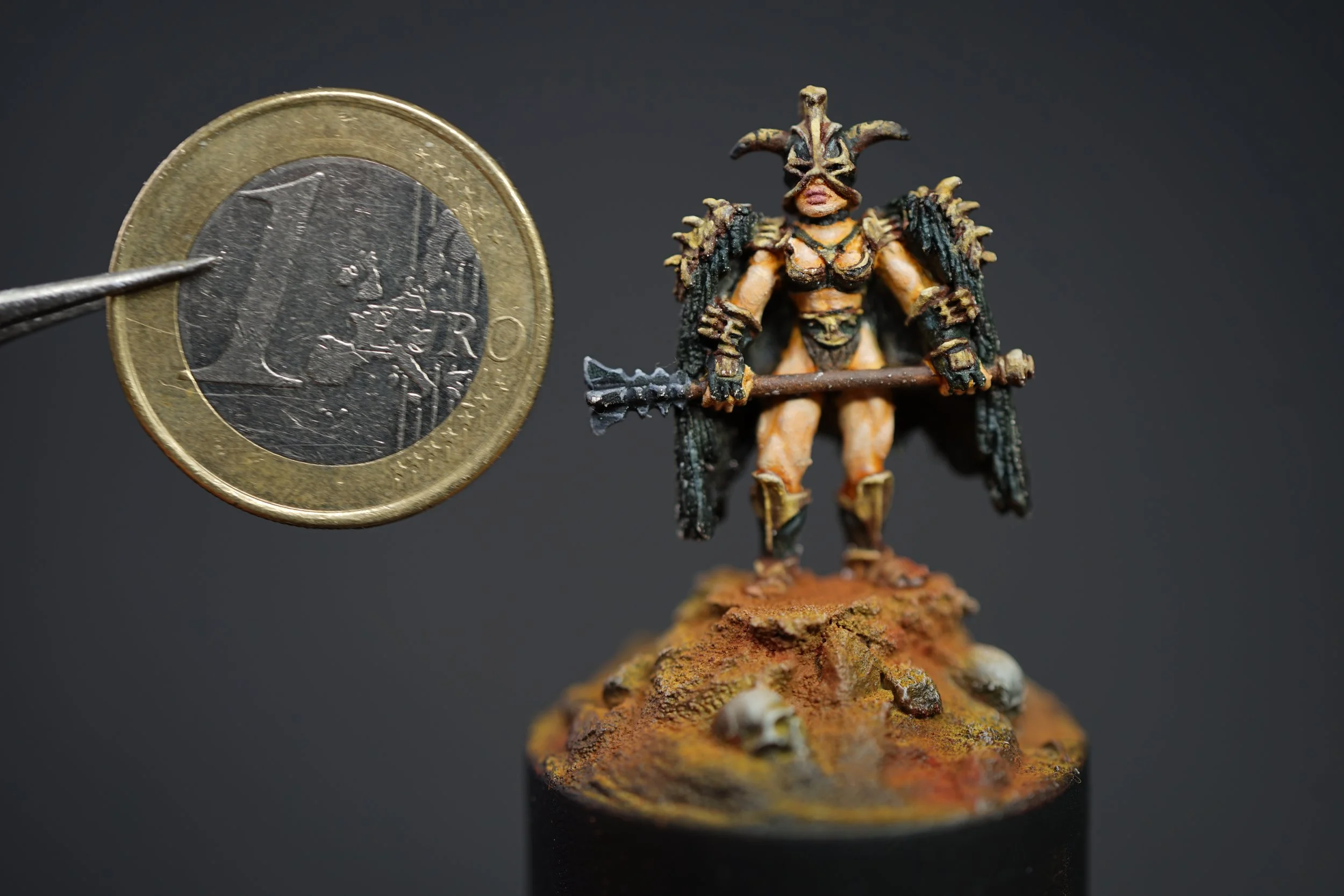 A miniatures figure of a warrior in armor, holding a mace, with a bird skull helmet and black cape, standing on a textured base with skulls, next to a one euro coin for size comparison, against a black background.