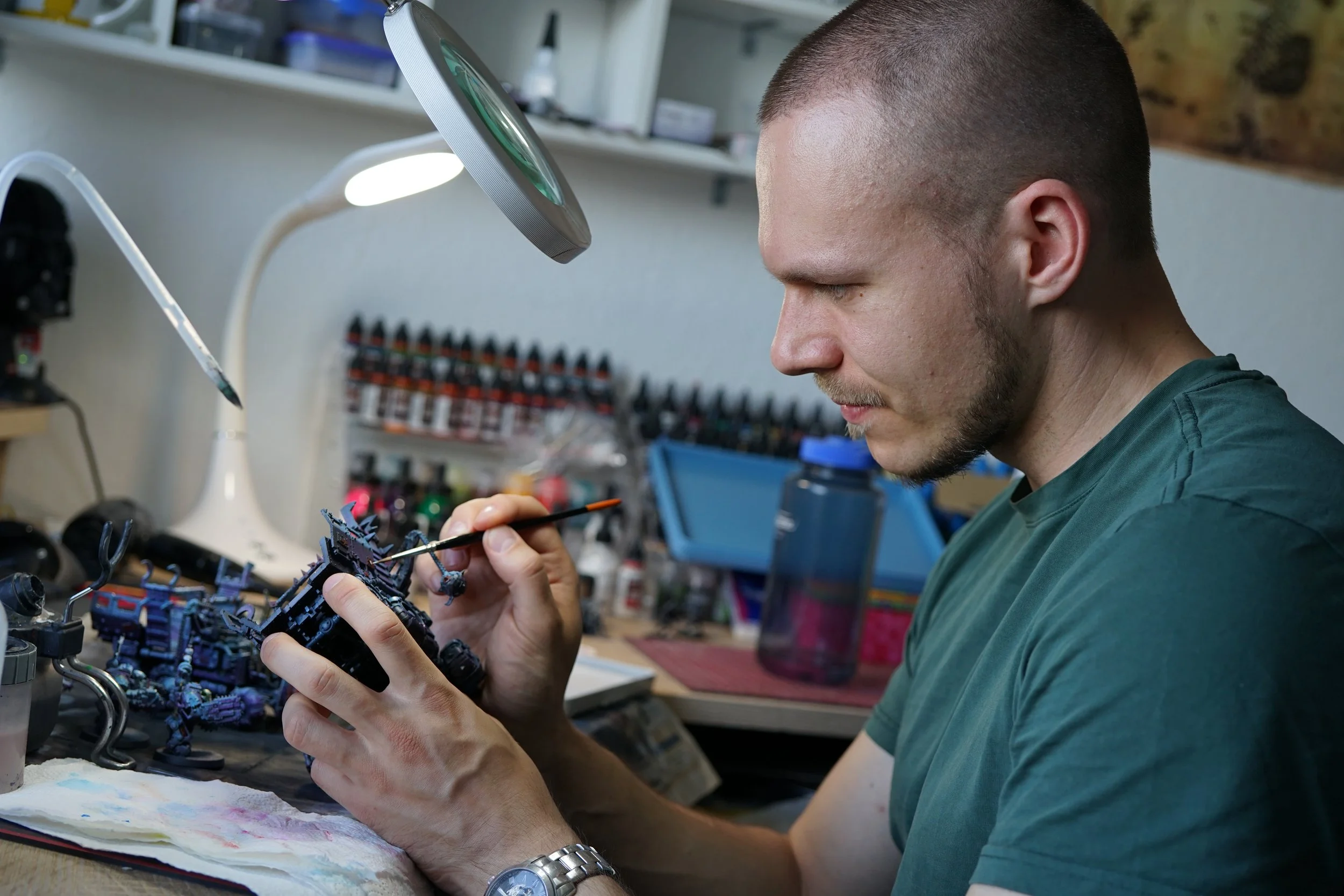 A man working on a miniature model at a workstation, using a fine brush under a desk lamp, with shelves of paint bottles in the background.
