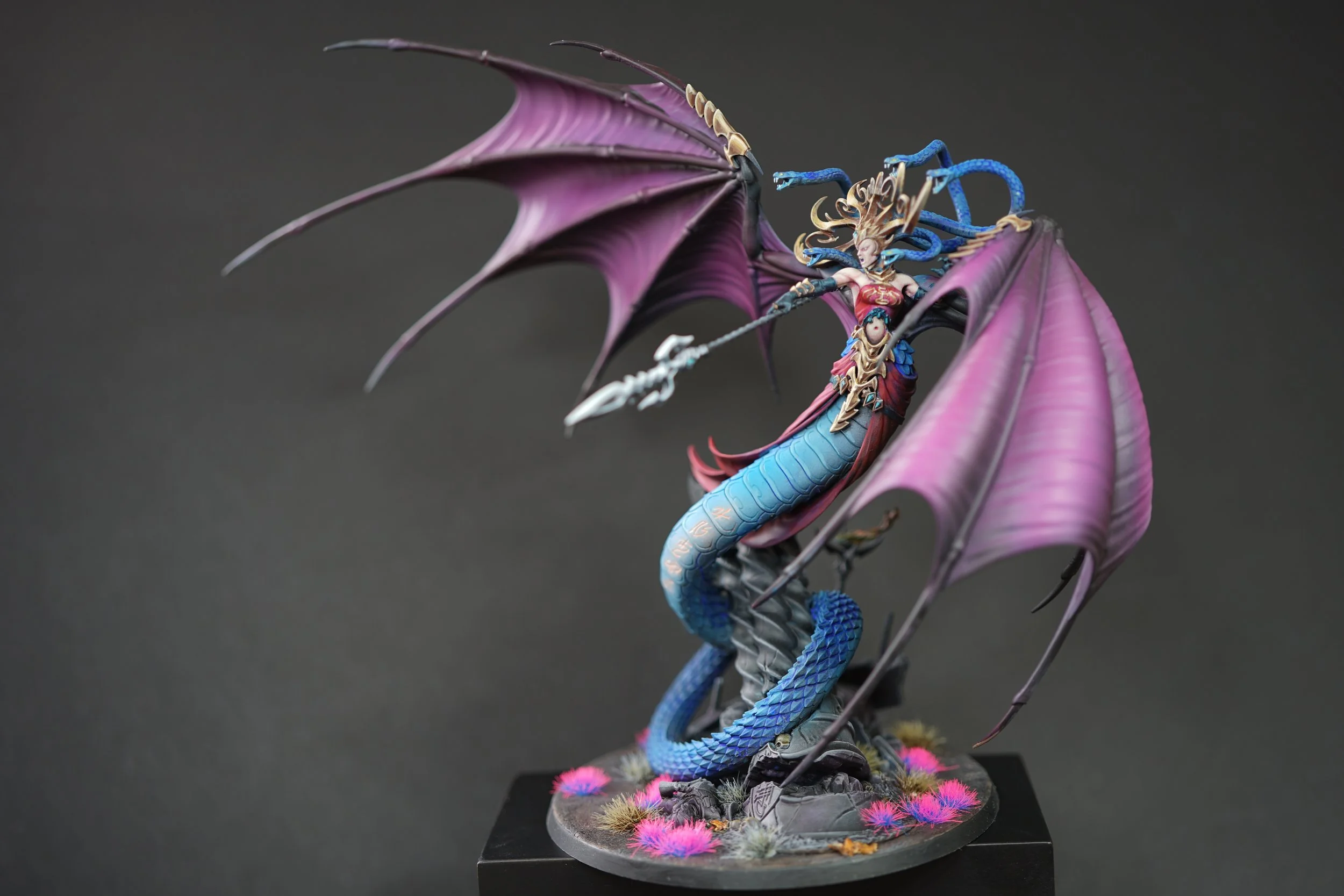 A detailed fantasy figurine of Morathi in her snake form from Warhammer Age of Sigmar with purple wings, blue hair, and a spear, standing on a rocky base with pink and yellow plants.