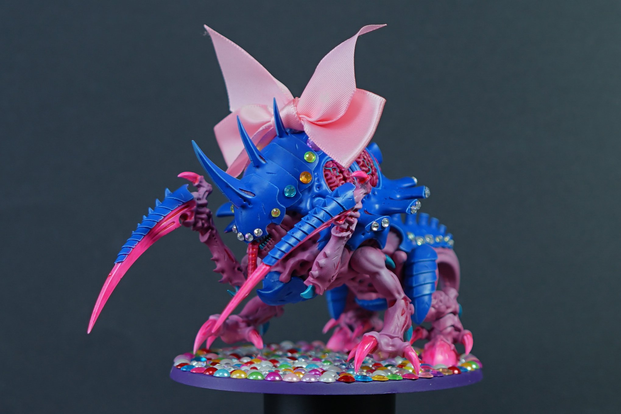 Colorful model of a Tyranid Maleceptor from Warhammer 40k with pink and blue colors, decorated with gemstones, a pink ribbon bow on its back, and multiple spiked armor plates, standing on a base covered with multicolored beads.