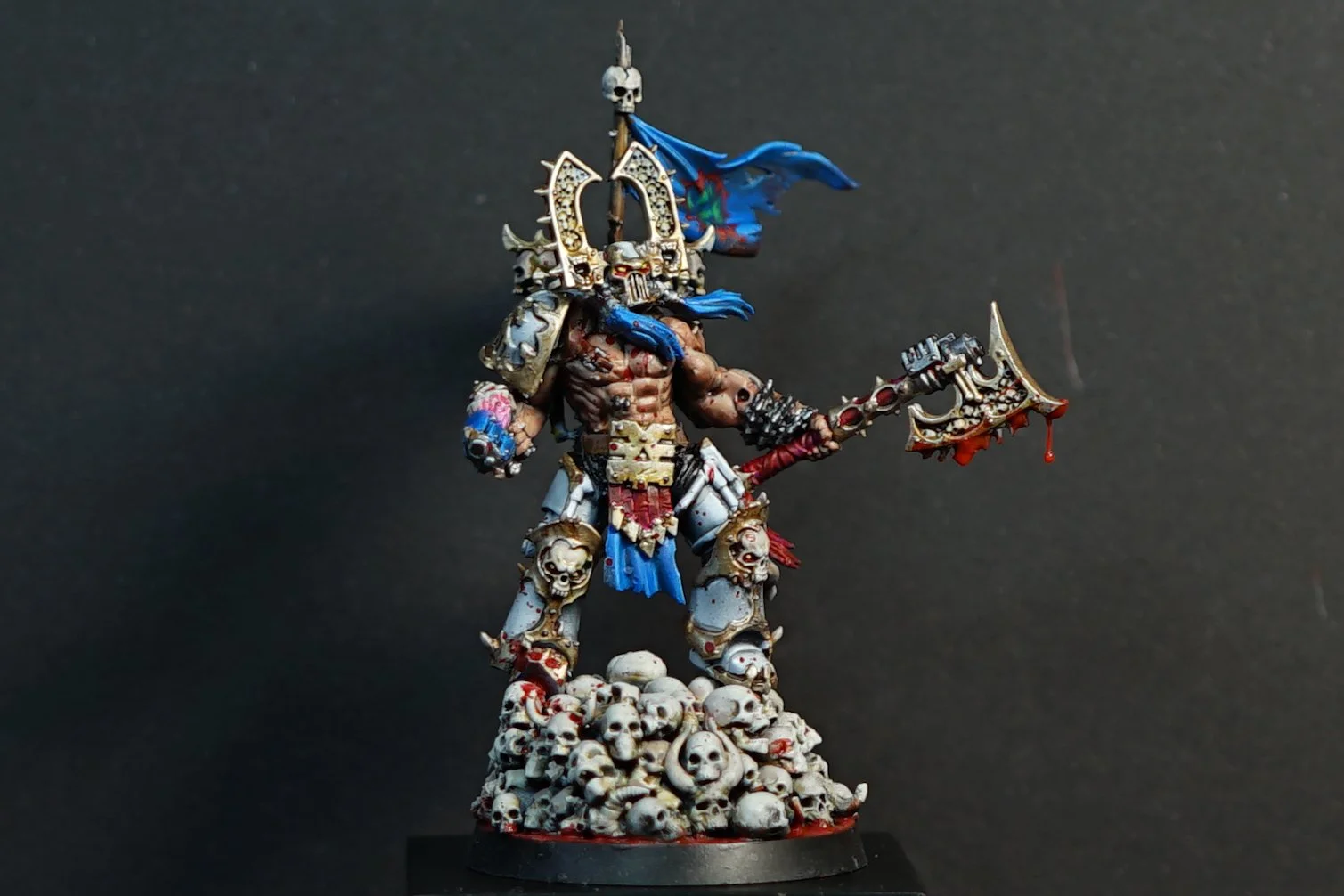 A painted and custom converted miniature of Khârn the Betrayer from Warhammer 40k on a base of skulls, wielding a spiked axe and wearing armor with skull motifs headdress, against a dark background.