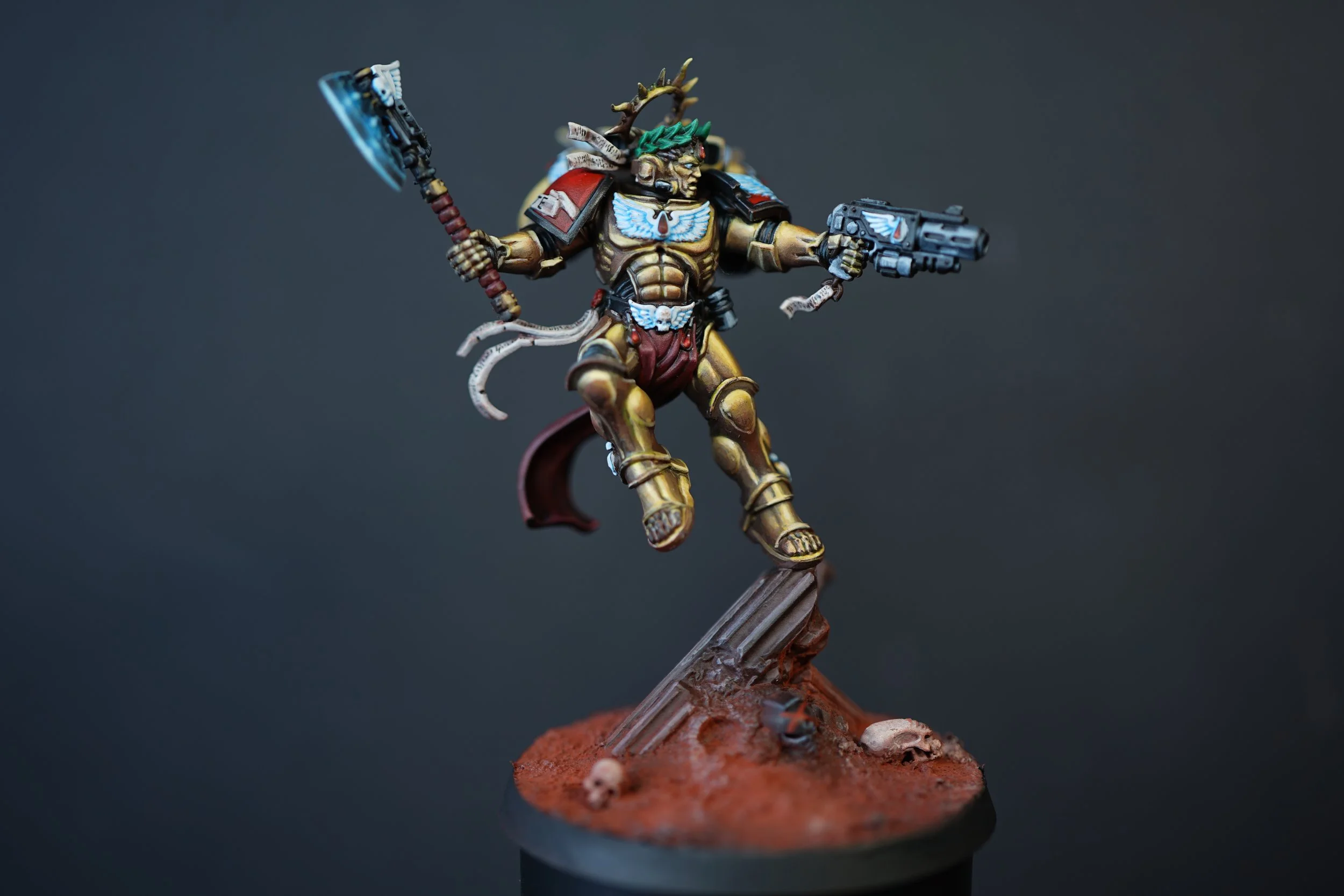 Miniature figurine of the Blood Angels chapter Master Dante from Warhammer 40k in his golden armor, holding a glowing axe and a gun, standing on a decorated base with skulls and bones, against a plain dark background.