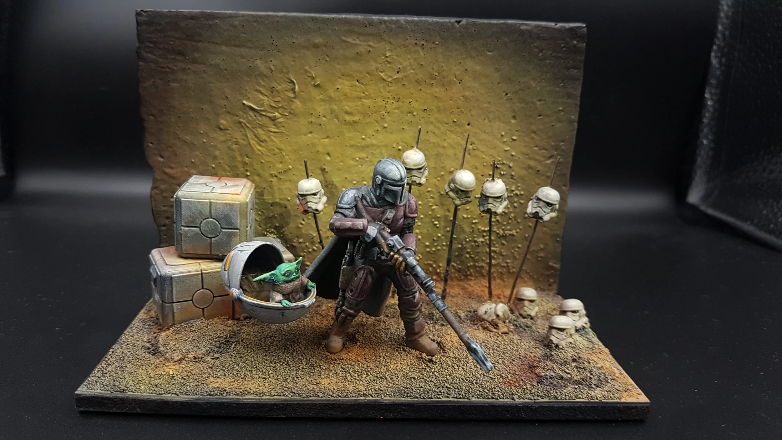 Miniature figurine scene featuring a Mandalorian holding a rifle, Baby Yoda in a pod, and stormtrooper helmets on sticks, set against a textured yellow background.
