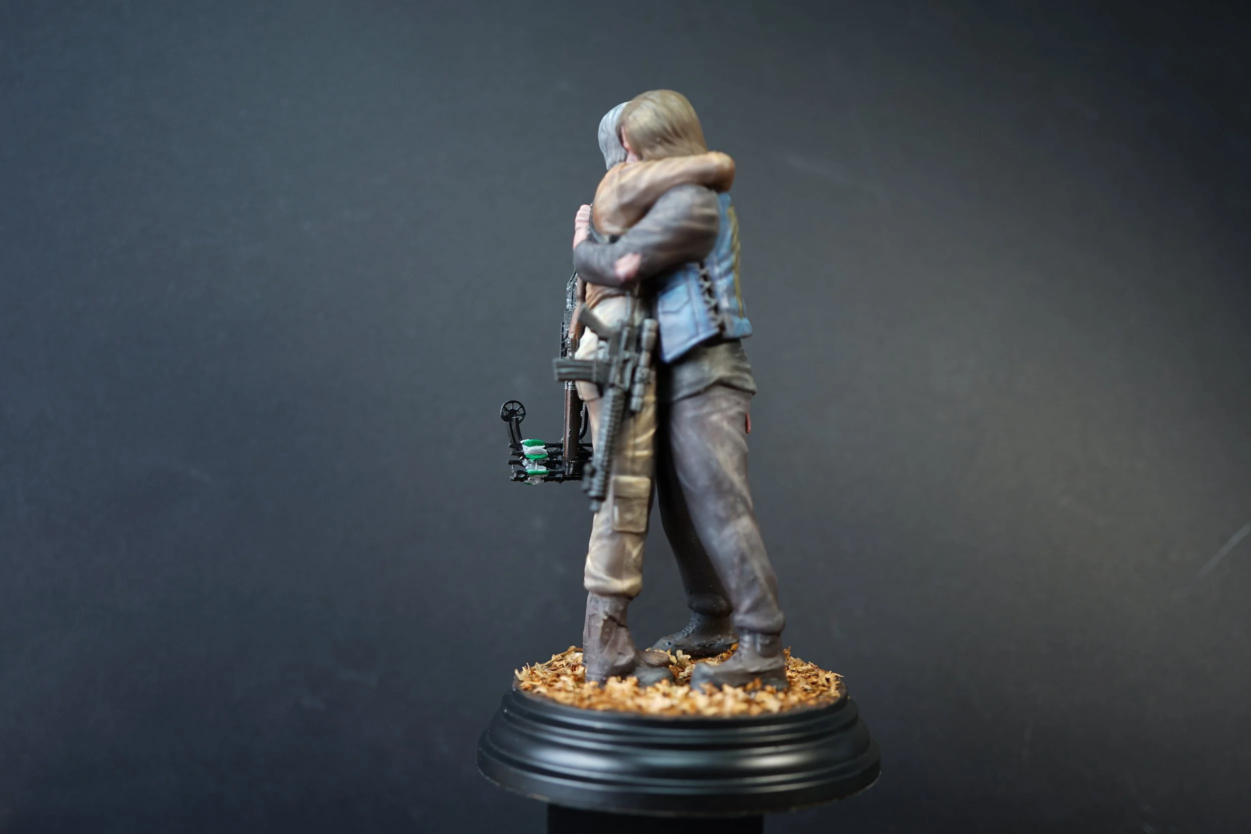 Miniature sculpture of Daryl and Carol from The Walking Dead series hugging, set on a round base with leaves, against a dark background.