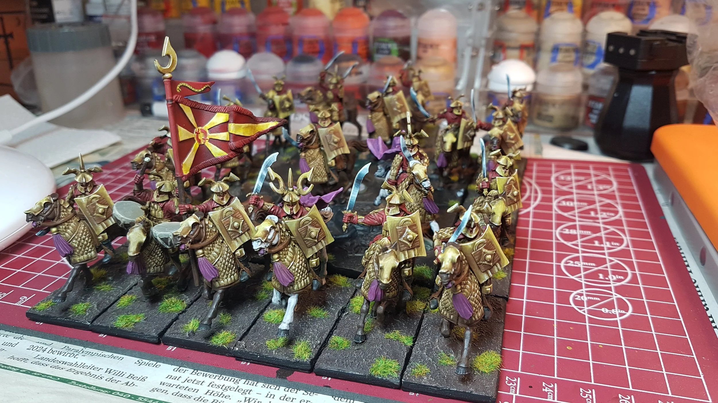 Miniature painted Easterlings from The Lord of the Rings on bases, mounted on horseback, holding shields and swords, with a banner, placed on a cutting mat with paint supplies in the background.
